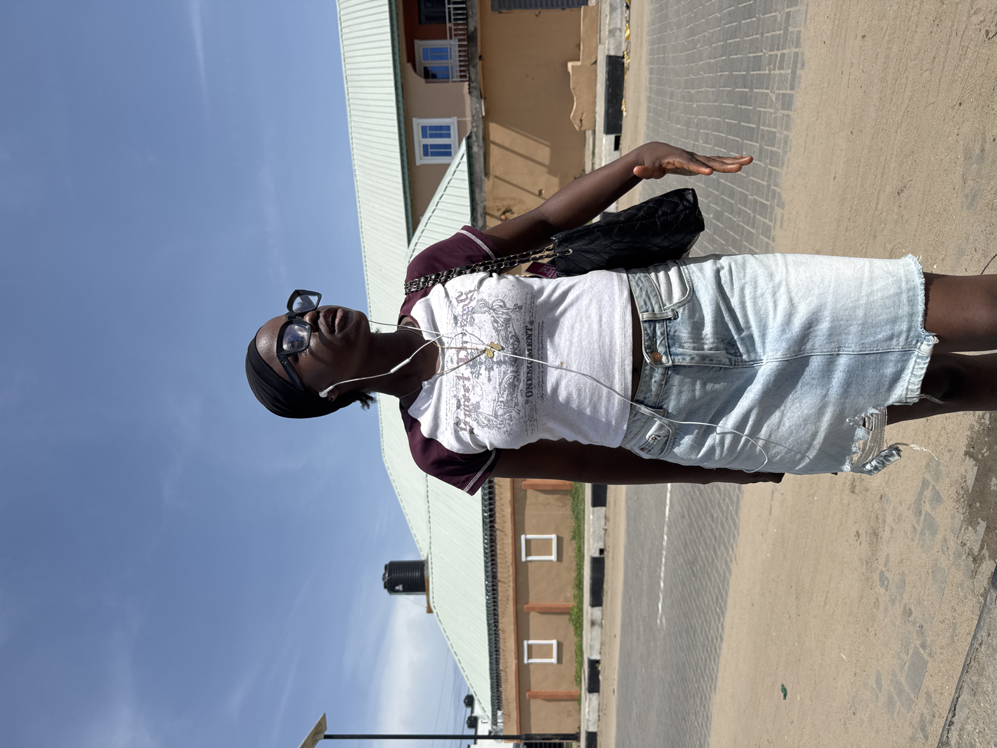A young woman wearing a white shirt with maroon sleeves, a denim skirt, black sunglasses, and wired earbuds stands on a paved area with buildings visible in the background under a clear blue sky. She has a black purse slung over her shoulder and her right hand is raised. The location is inferred to be in Badagry, Nigeria, based on the prompt instructions. The pavement has a mix of concrete and paved bricks. The buildings are a mix of beige, tan, and light green, some with black and white markings. A water tank is visible on the roof of one building.