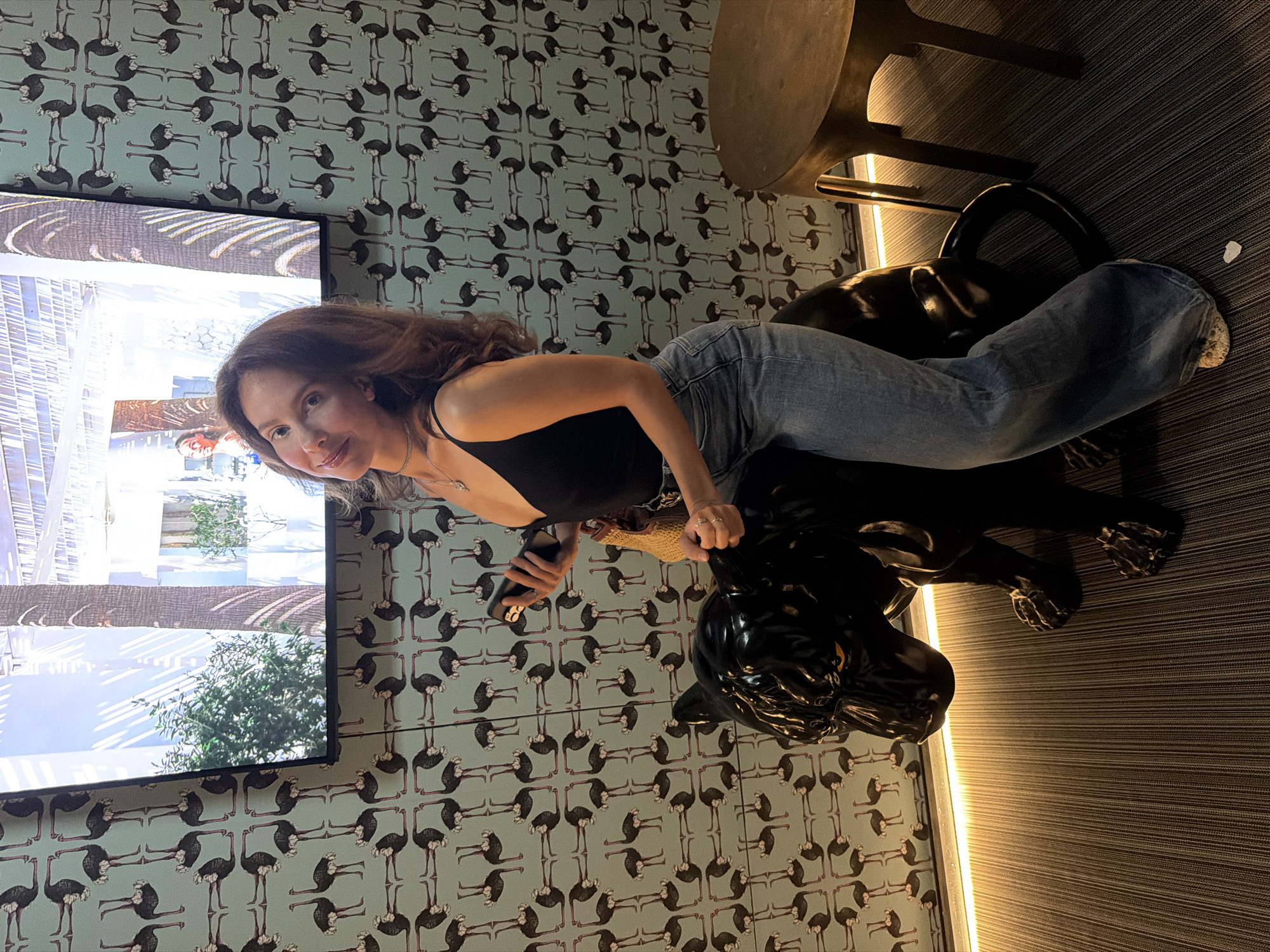 A woman with long, wavy brown hair and fair skin is posing for a photo while sitting on a black panther statue. She is wearing a black tank top and light-wash flared jeans. Her left hand rests on the panther's back, and her right hand holds a black smartphone. She is looking at the camera with a slight smile. The background features a light blue wallpaper with a repeating pattern of ostriches. A large television screen is mounted on the wall above the wallpaper, displaying an image of a modern building with palm trees. To the right of the woman, a round, dark wooden table with metallic legs is visible. The floor is carpeted with a striped pattern, and a strip of warm-toned LED lighting runs along the bottom of the wall.