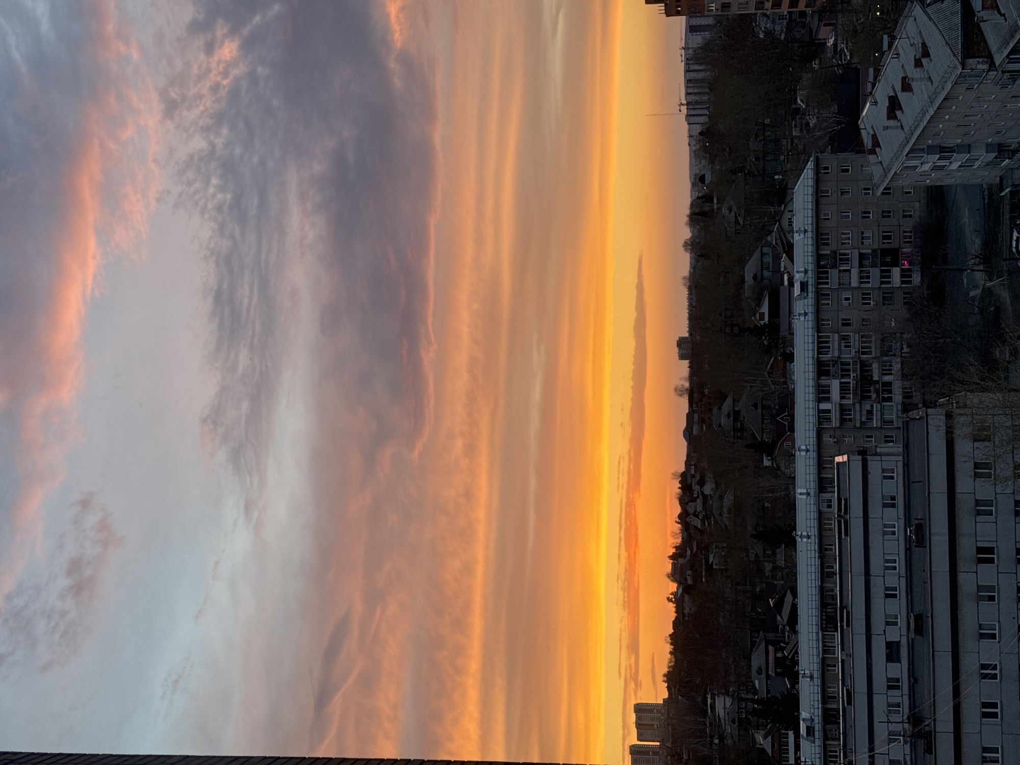 The image depicts a sunset over the city of Krasnoyarsk, Russia. The sky is filled with orange and grey clouds. In the foreground, there are apartment buildings. The setting sun is at the horizon. There are small buildings beyond the apartment buildings.