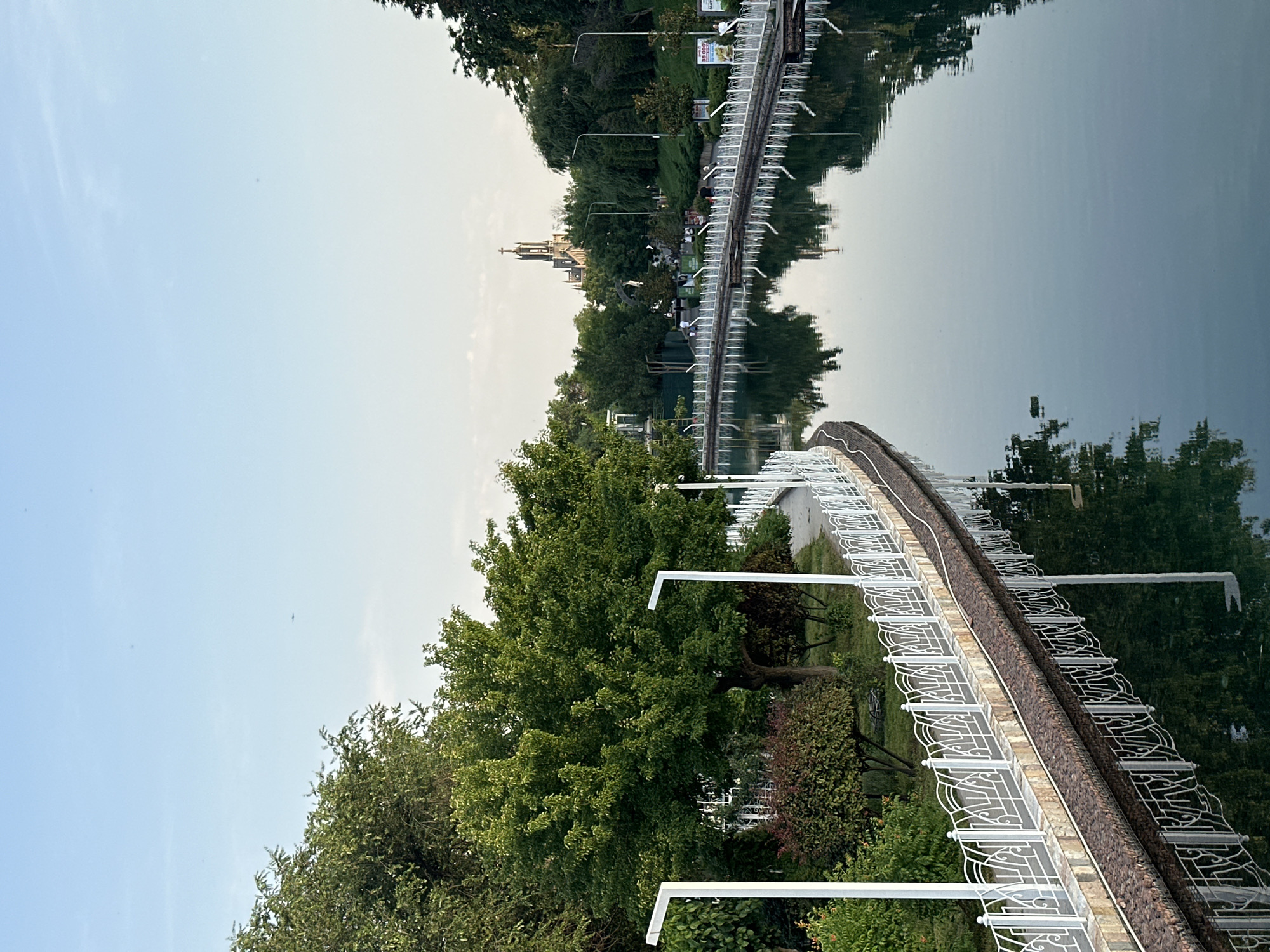 The image shows a view of a pond in Tashkent, Uzbekistan. A curved pedestrian bridge with white decorative railings and a brown brick pathway runs along the edge of the pond. The water surface reflects the bridge, trees, and a church tower in the distance. Lush green trees and bushes line the edge of the pond. Several lamp posts stand along the bridge. The sky is pale blue and hazy.