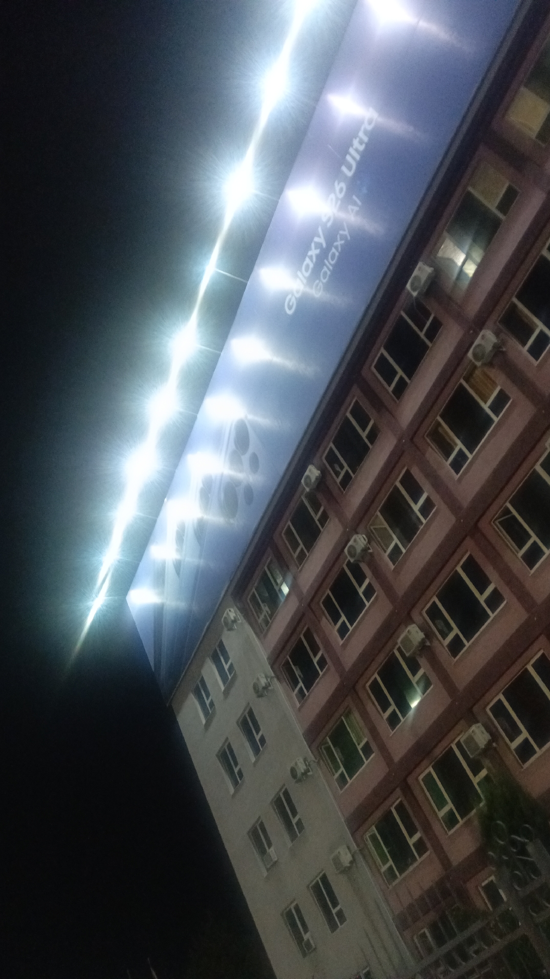 An outdoor, low-angle night shot shows a billboard advertising the "Galaxy S26 Ultra" and "Galaxy AI" positioned above a building with a pink facade. A row of bright lights illuminates the billboard from above, casting a glare. The building has multiple windows and air conditioning units visible. To the left of the pink building is a lighter-colored building. The sky is dark, and a fence is visible in the lower right corner.