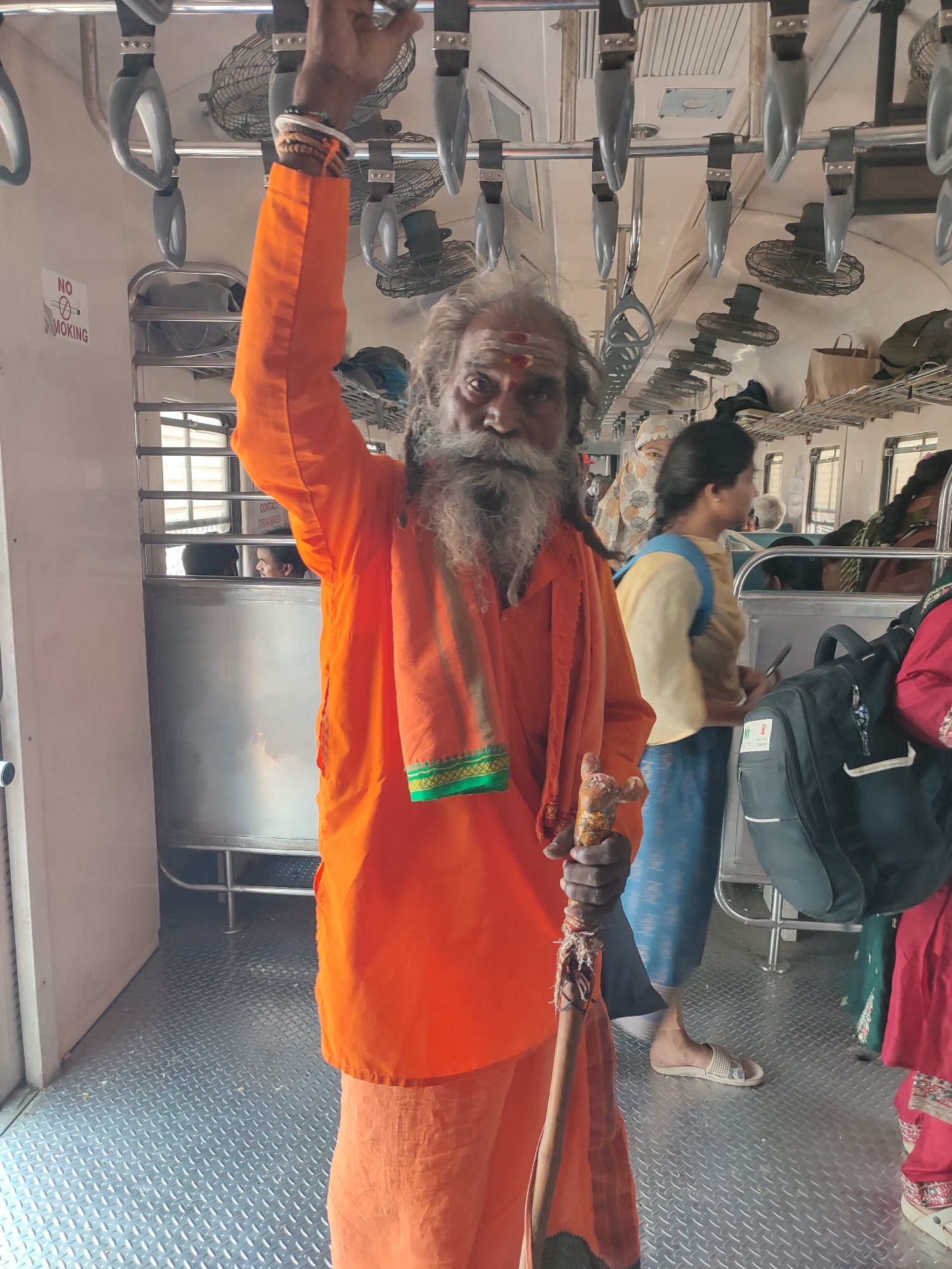 A full-shot image captures an elderly man, likely a sadhu or holy man, dressed in bright orange robes, standing inside a train carriage. He has a long, flowing white beard and dreadlocks, with a red and white tilak mark visible on his forehead. His right arm is extended upwards, grasping one of the many grey plastic overhead handles, while his left hand holds a wooden staff adorned with fabric. He wears a multi-strand bracelet on his right wrist.
The setting is a public transport train in India, indicated by the style of the carriage. The interior features rows of overhead metal luggage racks, ceiling-mounted fans, and numerous overhead hand-holds. The floor is a metallic, textured diamond plate. In the background, other passengers are visible; one woman in a yellow top and blue skirt stands near a black backpack, holding a phone. Further back, more passengers are seated or standing. A "NO SMOKING" sign with an angled cigarette icon is prominently displayed on the left wall of the carriage. Faintly visible on a window reflection are the words "CONTACT" and a partial phone number "7704 60453". The scene depicts a typical moment of travel on an Indian train, with the main subject seemingly posing for the camera.