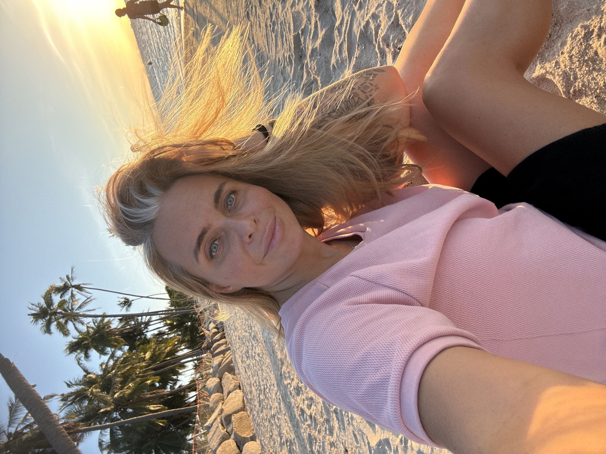 A woman with blonde hair and blue eyes, wearing a pink shirt and black shorts, is sitting on a sandy beach at sunset. She is looking at the camera with a slight smile. In the background, there are palm trees, rocks, and the ocean. A person is visible walking on the beach in the distance. The sky is a warm orange and yellow due to the sunset. The scene is likely Baan Tai, Thailand, given the presence of palm trees and the beach environment typical of the area.