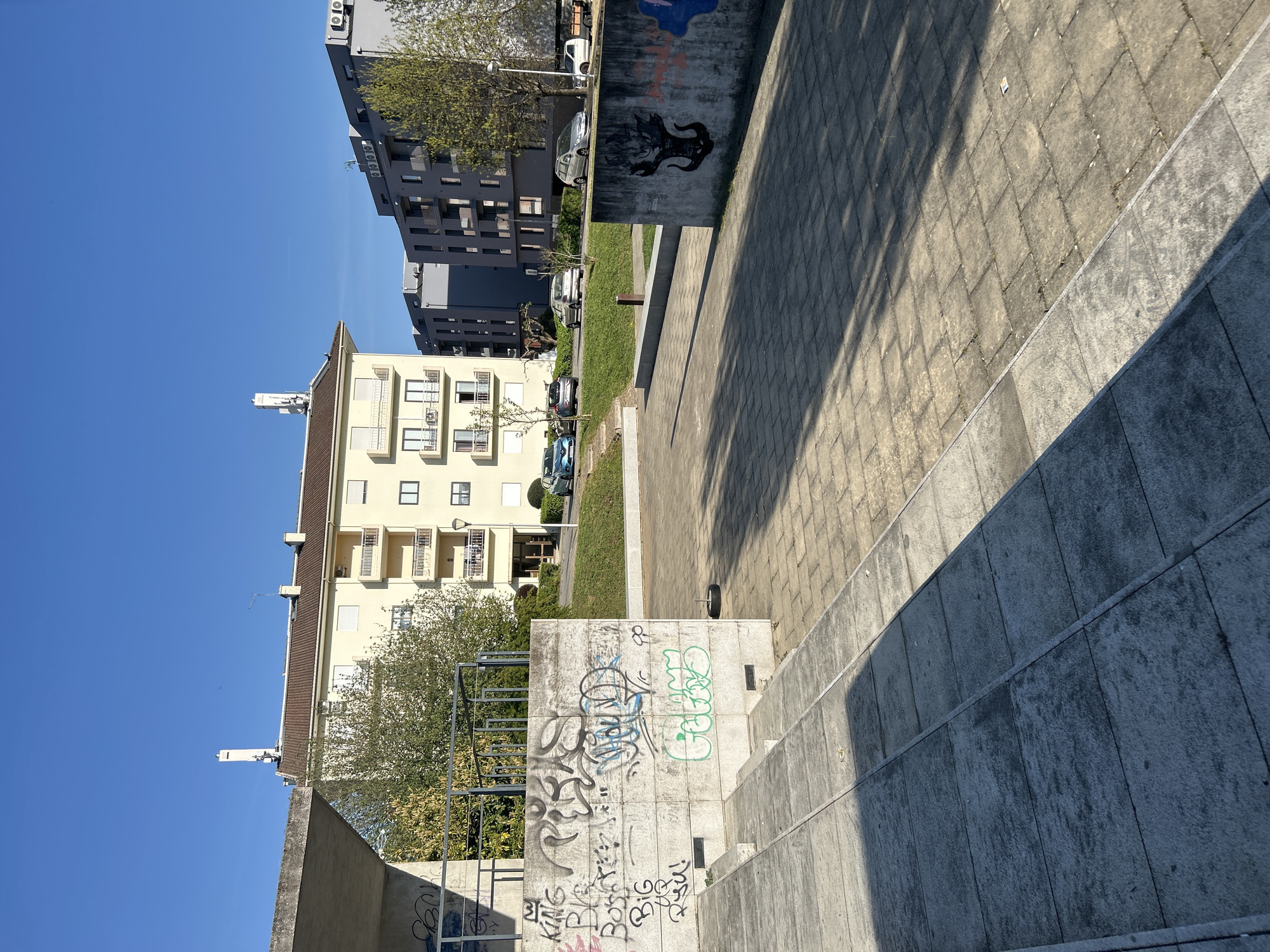 The image shows an outdoor urban scene in Vila Real, Portugal. A series of concrete steps lead down to a paved area. Walls covered in graffiti flank the steps. Behind the wall, there are buildings, trees, and parked cars. The sky is blue. There are no people visible.