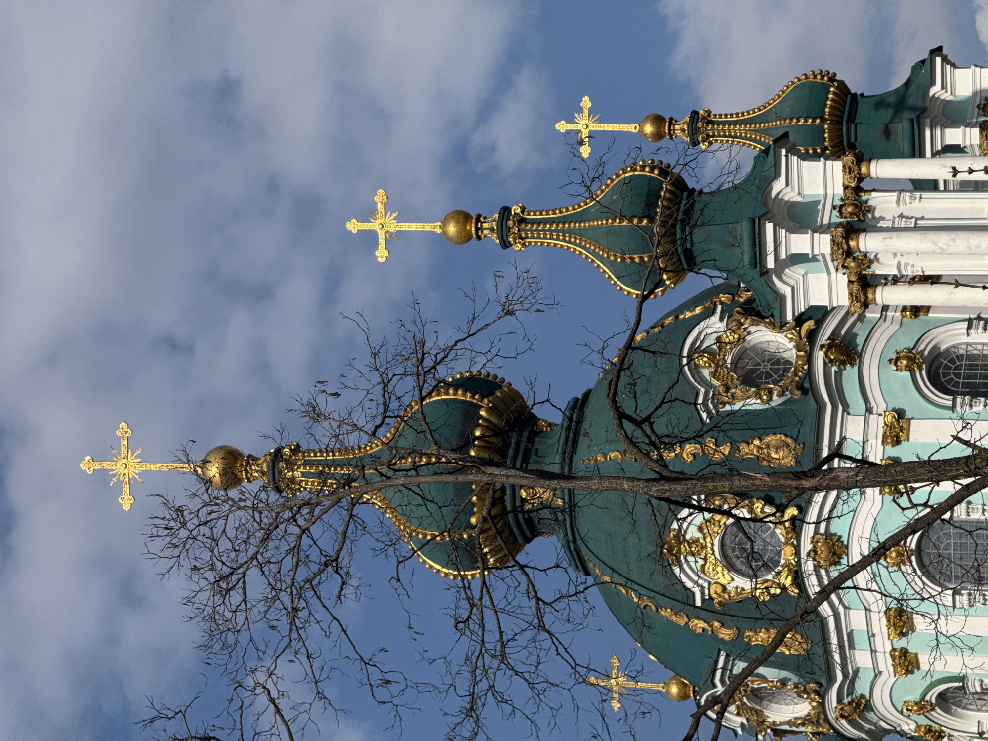 The image displays the upper sections of St. Andrew's Church, located in Kyiv, Ukraine. The architecture features multiple bulbous domes and spires, primarily teal-green with extensive gold ornamentation. A large central dome is visible, flanked by smaller, similarly styled spires. Each dome and spire is topped with a golden Orthodox cross; the prominent cross at the highest point features an ornate sunburst design. The visible facade sections below the domes are rendered in a light blue-green and white, also adorned with gold baroque-style detailing, including sculpted motifs and circular windows with grilles. The foreground contains the intricate network of bare, dark branches of a deciduous tree, partially obscuring the church structure. The background is a blue sky with scattered white cumulus clouds. No people or discernible actions are present.