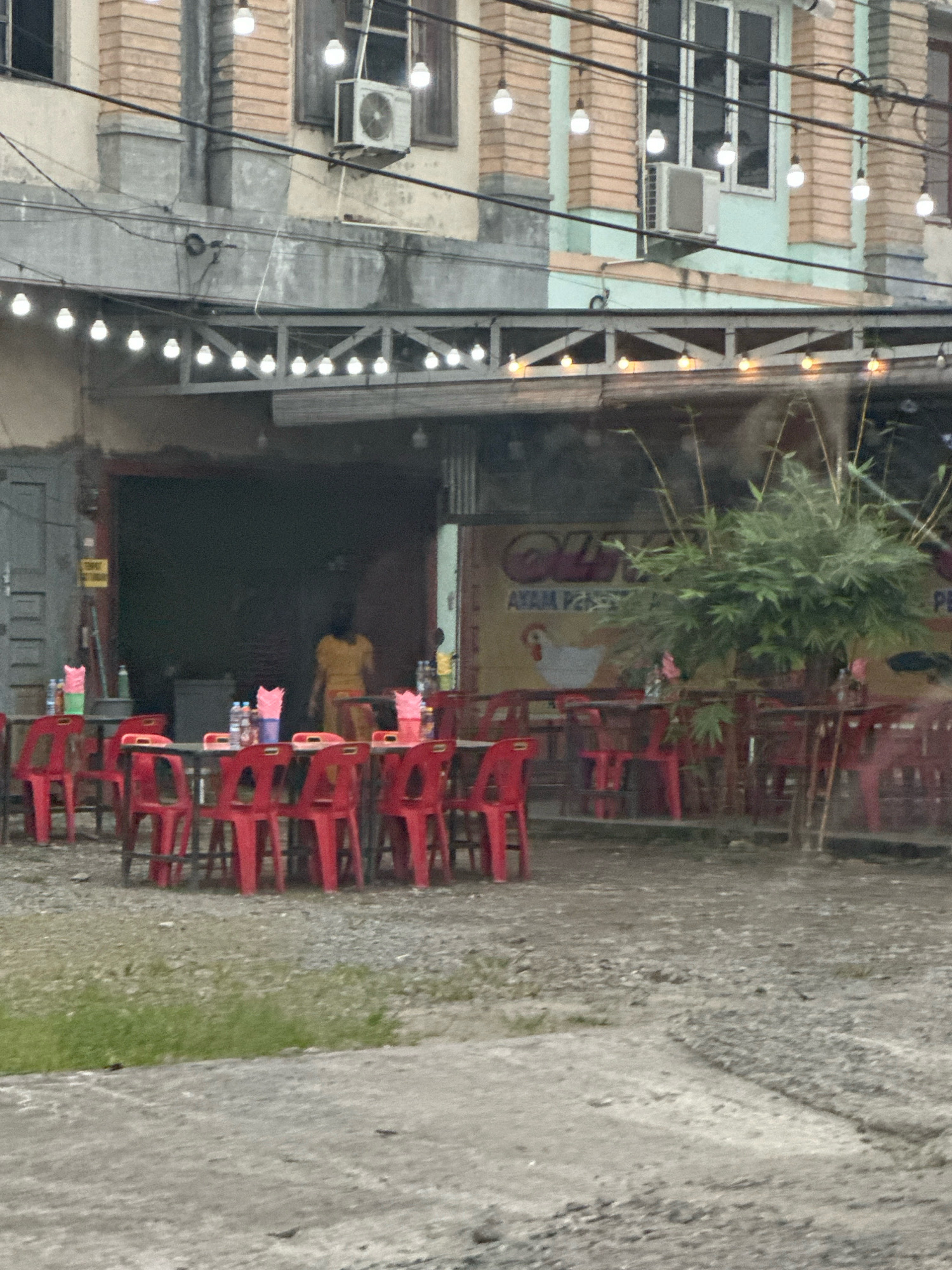 The image captures an outdoor seating area of a restaurant in the City of Binjai, Indonesia. Red plastic chairs and tables are arranged on a gravelly ground, with some grassy patches visible. In the background, a building with a light-colored facade and brick accents is seen. Strings of lights are strung across the top of the seating area. A woman in a yellow dress stands near the entrance of the establishment. A potted bamboo plant is positioned to the right, partially obscuring a yellow sign with text and an illustration of a chicken. The scene appears to be a casual dining setting, possibly a roadside eatery, given the simple furniture and open-air arrangement. The lighting suggests it might be daytime, though the overall light level is low, indicating potential overcast conditions or late afternoon.