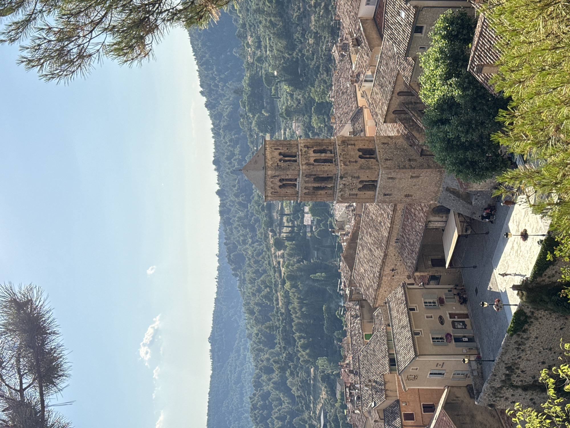 This is an aerial view of Moustiers-Sainte-Marie, France, a picturesque village nestled in the Alpes-de-Haute-Provence department. The scene is dominated by the iconic bell tower of the Notre-Dame de Beauvoir church, which stands tall amidst a cluster of terracotta-tiled roofs. The village is set against a backdrop of lush green hills, dotted with trees and houses, suggesting a rural or semi-rural mountainous setting.
In the foreground, a stone wall encloses a sun-drenched plaza paved with light-colored cobblestones. Several people are visible in the plaza, some walking and others seated outside a building that appears to be a cafe or restaurant, judging by the tables and chairs. Streetlights with hanging flower baskets line the plaza.
The architecture of the village features stone buildings with tile roofs, characteristic of the region. The bell tower itself is a four-tiered structure with arched openings on each level and a pyramidal roof. The surrounding buildings are a mix of residential and commercial spaces, with shops on the ground floor of some.
The sky is clear blue with a few wispy clouds, and sunlight casts shadows, indicating it is daytime, likely mid-morning or afternoon. Pine branches in the upper and lower parts of the frame frame the view, adding a natural element to the man-made landscape. The overall impression is one of tranquility and a preserved historic European village.
