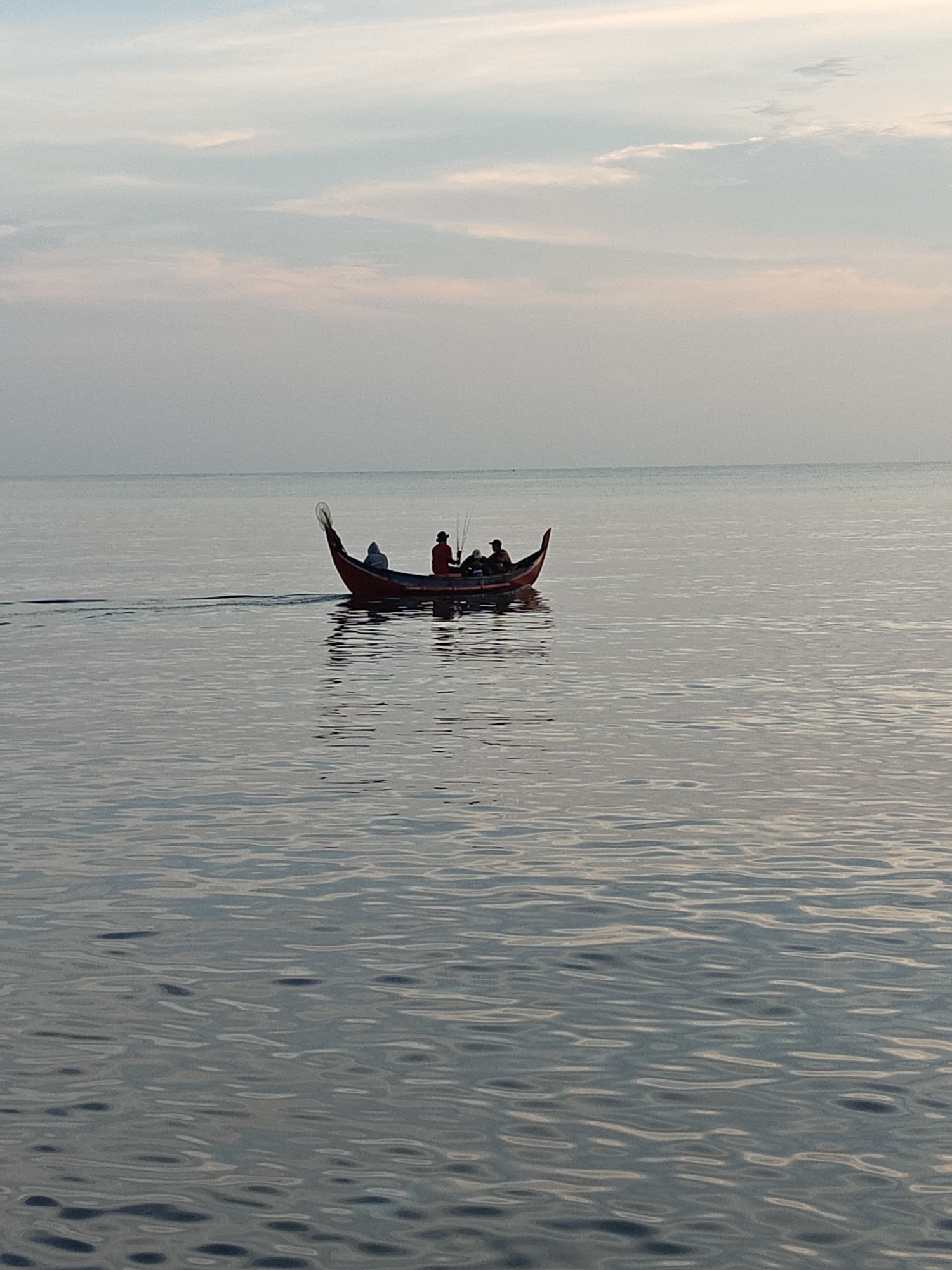 A serene seascape at dawn or dusk shows three silhouetted figures in a long, narrow, red-and-dark-toned traditional boat, moving across calm waters. The sea surface is rippled, reflecting the soft light from the sky, which is pale with hints of pink. The boat leaves a faint wake behind it, indicating movement. The scene is likely situated off the coast of Lhokseumawe, Indonesia, judging by the style of boat.