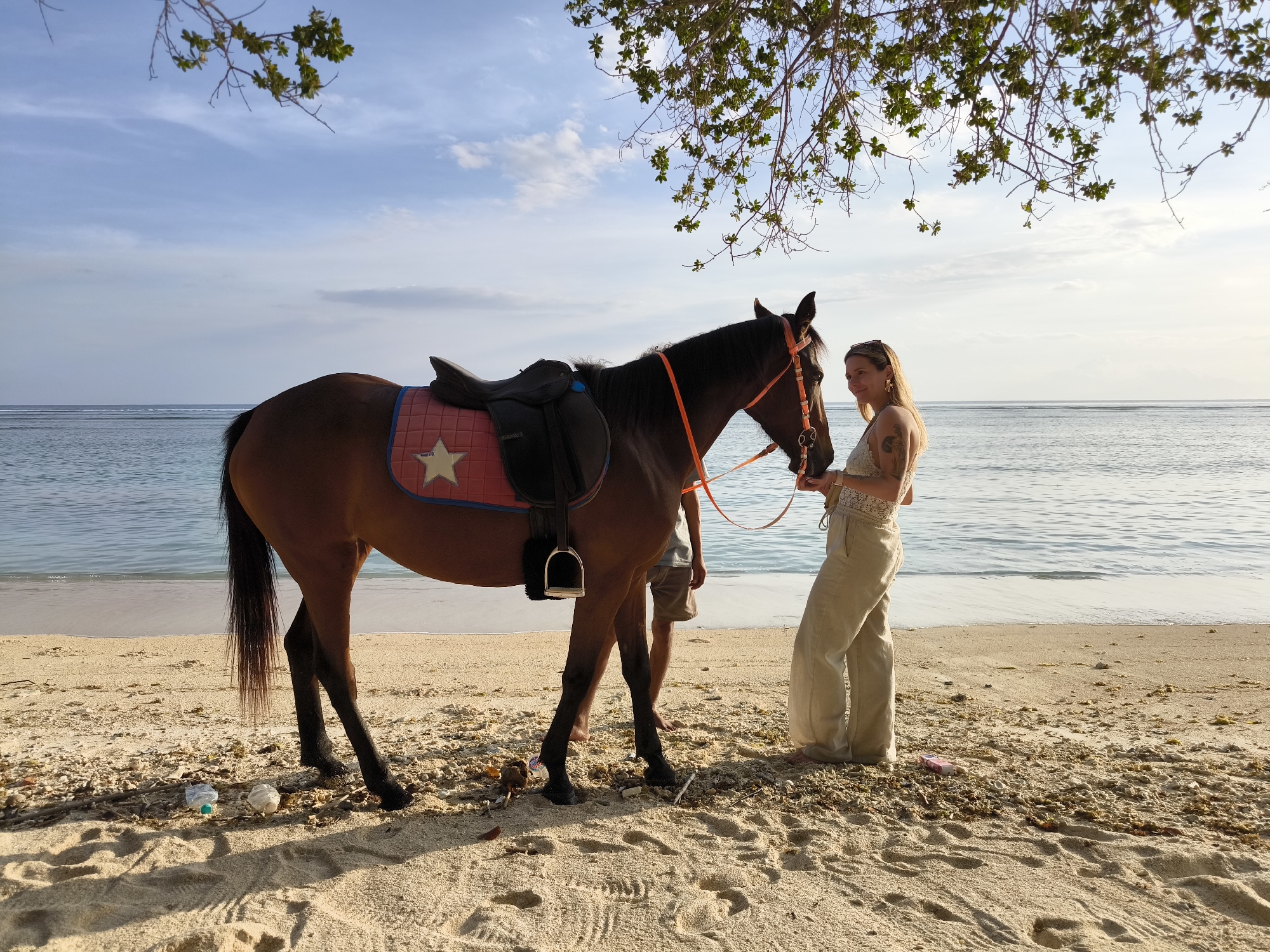 A woman in loose tan clothing stands on a beach near a brown horse with a saddle. The horse is held by an orange bridle. Another person stands behind the horse. The background is a beach in Unknown, Indonesia, with blue water. There are trees and trash on the sand.