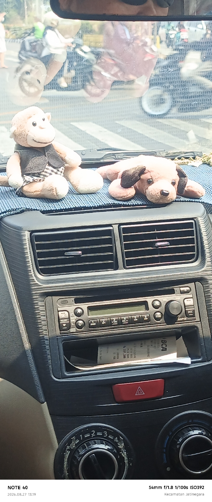 In the foreground of the image, the dashboard of a vehicle is visible. Two plush toys, a monkey and a dog, are placed on the dashboard. The monkey is wearing a gray vest over a checkered shirt and shorts, while the dog is lying down. A car stereo system with buttons and a display screen is mounted in the center of the dashboard. Below the stereo, there is a storage compartment containing a receipt. The background shows a blurred reflection of a street scene, with vehicles, including motorcycles and cars, and people. The timestamp at the bottom indicates the photo was taken on August 27, 2026, at 13:19 in Kecamatan Jatinegara, East Jakarta, Indonesia.