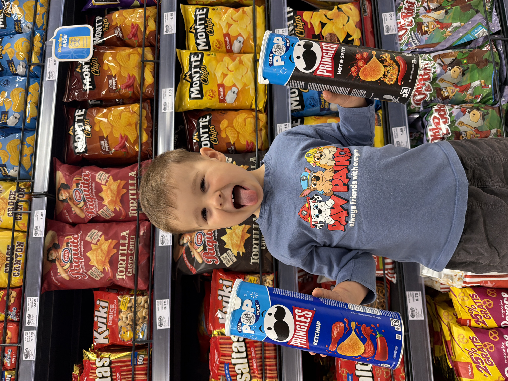 A young boy with blond hair, wearing a blue long-sleeved t-shirt adorned with "PAW PATROL always friends with everyone" characters, stands in a brightly lit supermarket aisle. He is facing the camera with a playful, excited expression, mouth wide open and tongue sticking out. He holds two Pringles cans, one in each hand, against a backdrop of snack-filled shelves.
In his right hand, the boy holds a black Pringles can of "HOT & SPICY" flavor, featuring an illustration of a chili pepper and a chip. Visible text includes "SCAN POP PLAY" and "Гострий та пряний смак" (Hot and spicy taste). In his left hand, he holds a blue Pringles can of "KETCHUP" flavor, depicted with ketchup pouring onto a chip. This can also shows "SCAN POP PLAY" and "СМАК КЕТЧУПУ" (Ketchup taste) and "КАРТОПЛЯНІ ЗІ СМАКОМ" (Potato chips with flavor).
The shelves around him are well-stocked with various snack items. To his left, red bags of "PATA Paquita TORTILLA Gusto Chili" are prominent, alongside "Kviki RIBICE S MAKOM" and "saltas STAPICI-STICKS". Price tags, some displaying the "MEGA" logo and values like "0.75", "1.15", and "0.65", are attached to the shelves. To his right, yellow bags of "MONTTTE Chips" in "Classic" and "Chilli Pepper" flavors are visible, along with other chip brands. A blue sign with "Kupujmo NAŠE!" (Buy Local!) is positioned on a higher shelf. The prices suggest a European currency, consistent with the context of Budva, Montenegro. The scene captures a joyful moment of a child selecting snacks during a shopping trip.