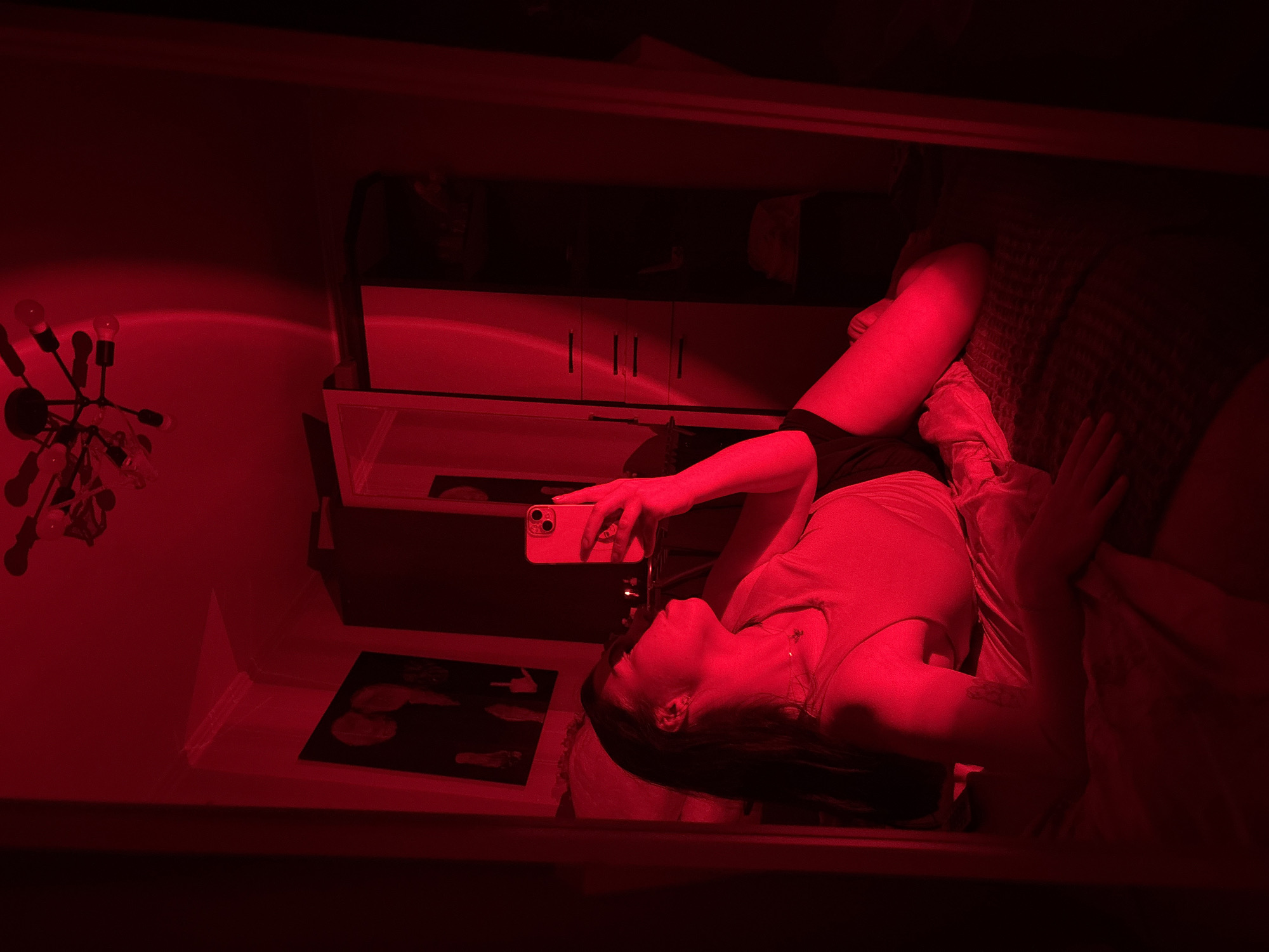 The image is a red-toned indoor reflection. A person is lying on a cushioned surface, holding a cell phone in their hand, presumably taking a picture in the mirror. Reflected in the mirror are various objects: a light fixture with multiple bulbs, a cabinet with drawers and doors, and a framed picture on a lower surface. The reflection suggests the photo was taken in a domestic interior. There is no information indicating the city of Perm, Russia.