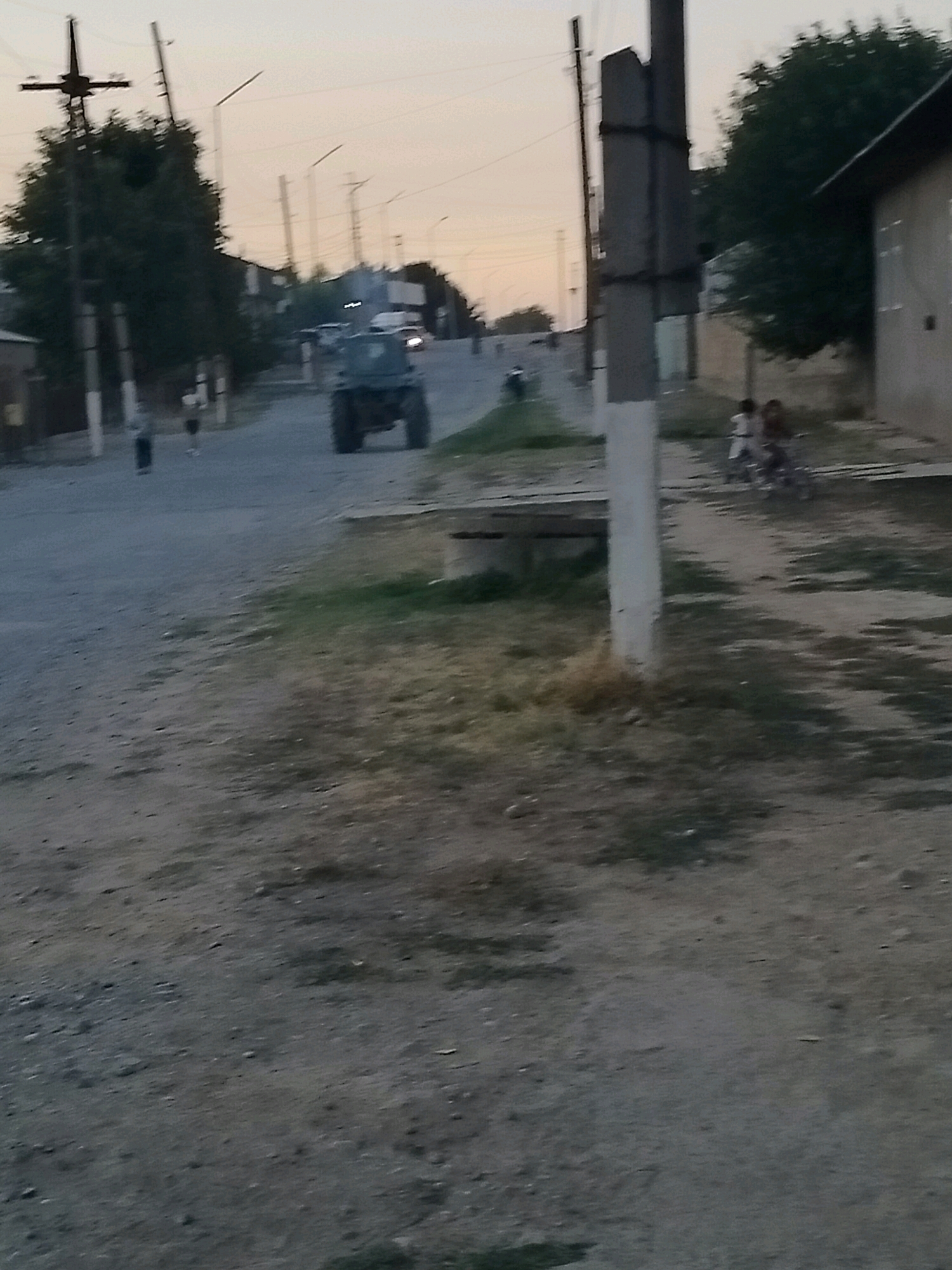 The image depicts a street scene in the city of Kölkent, Kazakhstan, likely during the late afternoon or early evening given the soft, diffused light. A dark-colored tractor is the most prominent subject, positioned in the center of the paved road, moving away from the viewer. Several pedestrians are visible on the left side of the road. Two individuals, appearing to be children, are on bicycles or a similar wheeled vehicle on the right side of the road, near a building. The environment features residential buildings, utility poles with wires, and trees. The road surface is paved, with gravel and dirt on the sides, suggesting a semi-urban or developing area. The overall atmosphere is subdued and calm.