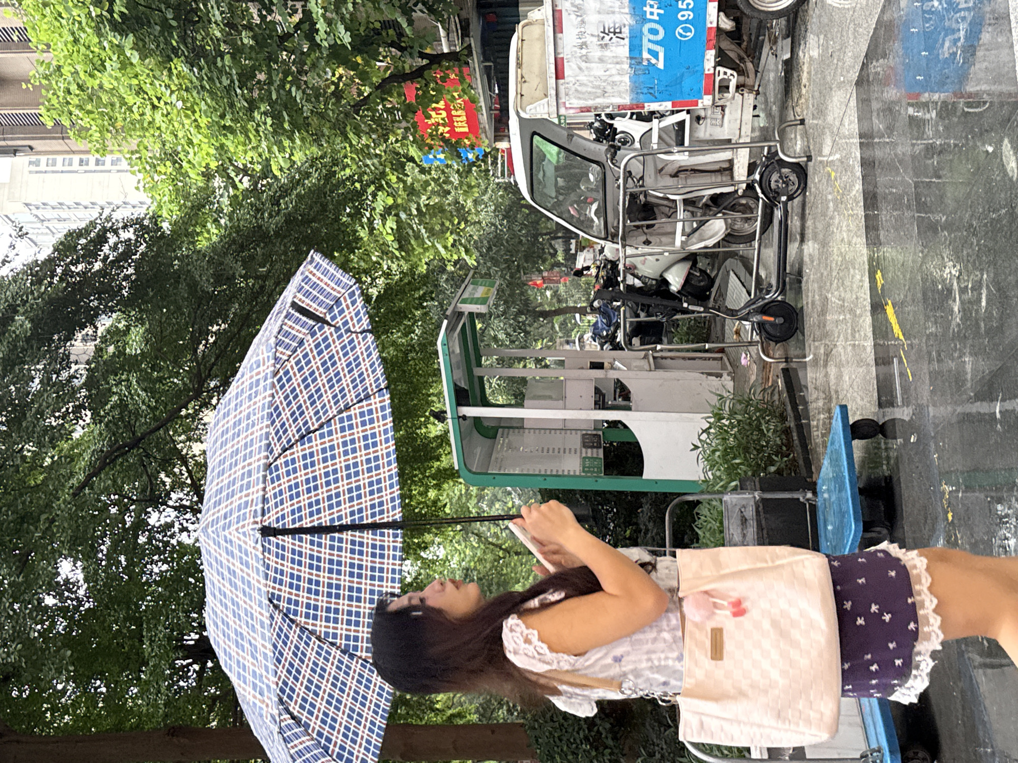 A young Asian woman is walking under a blue and white plaid umbrella on a wet city street in Haizhu District, China. She is holding a white smartphone in her right hand and carrying a light pink checkered tote bag on her left shoulder. The woman is wearing a white lace-trimmed top and a dark purple skirt with white floral patterns. In the background, there is a green and white charging station for electric scooters, several parked scooters, and a white three-wheeled delivery vehicle with blue and red signage. Trees with lush green foliage line the street, and tall buildings are visible in the distance. The ground is wet and reflective, indicating recent rain.