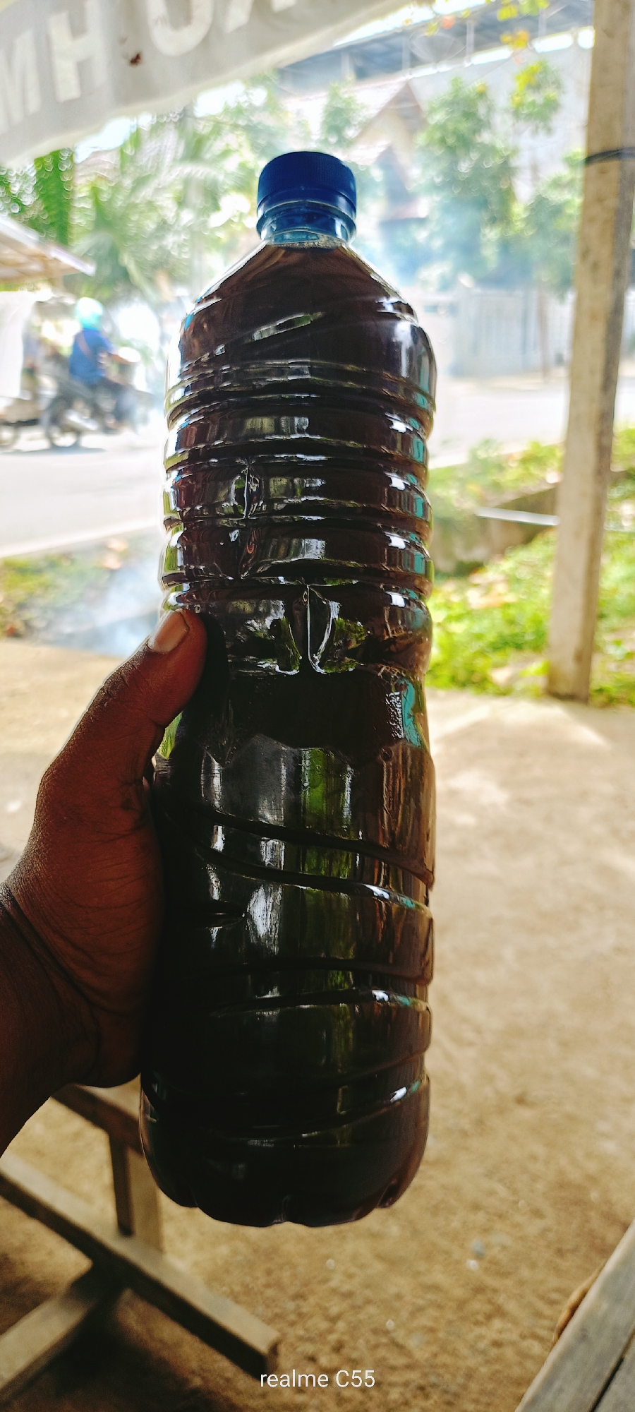 A dark brown liquid fills a plastic bottle with a blue cap, held in a person's hand. The bottle has a ribbed texture. The background is blurred, showing greenery and some indistinct buildings, suggesting an outdoor setting. A motorcycle with a rider wearing a blue shirt and helmet is visible in the distance on the left. The overall impression is that of a casual, everyday scene, possibly in a market or roadside stall in Lhokseumawe, Indonesia.