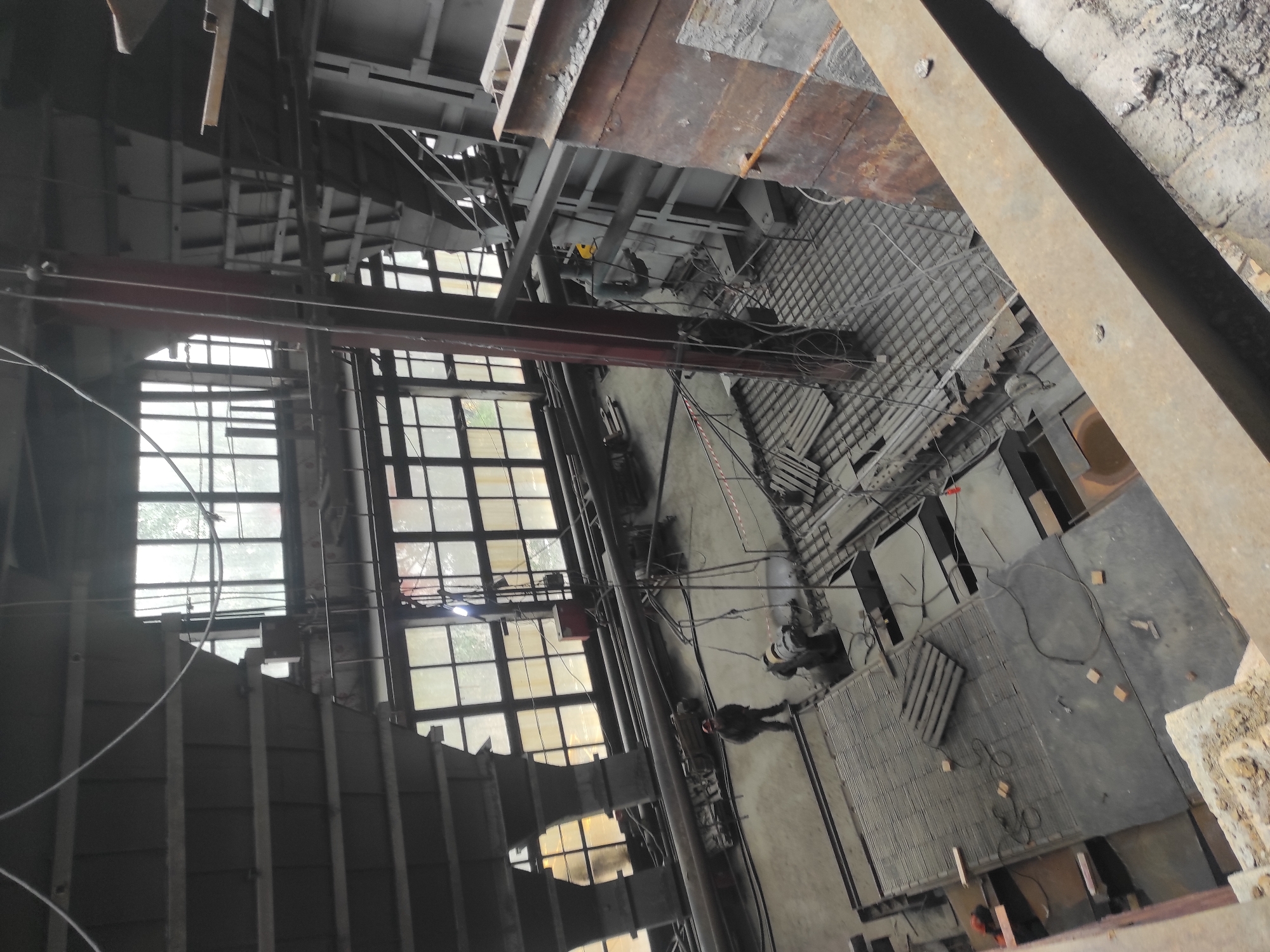 This image shows an interior view of a large industrial facility, likely under construction or renovation, in Kyzyl, Kazakhstan. The space is characterized by its high ceilings, large windows with multiple panes, and exposed structural elements such as steel beams and columns. Several industrial materials and equipment are visible, including rebar on the floor, pipes, and what appear to be machinery or large components.
In the foreground, there is a metal beam and concrete debris. Moving deeper into the facility, a person wearing a hard hat and work clothing stands in the center of the main floor. They are positioned near some machinery and electrical cables. Various sections of the floor are at different levels, suggesting a multi-tiered structure. Wooden pallets are stacked on some platforms.
The lighting in the facility appears to be natural, coming from the large windows, and supplemented by artificial lights, one of which is visible near the ceiling. The overall atmosphere is that of an active construction site.