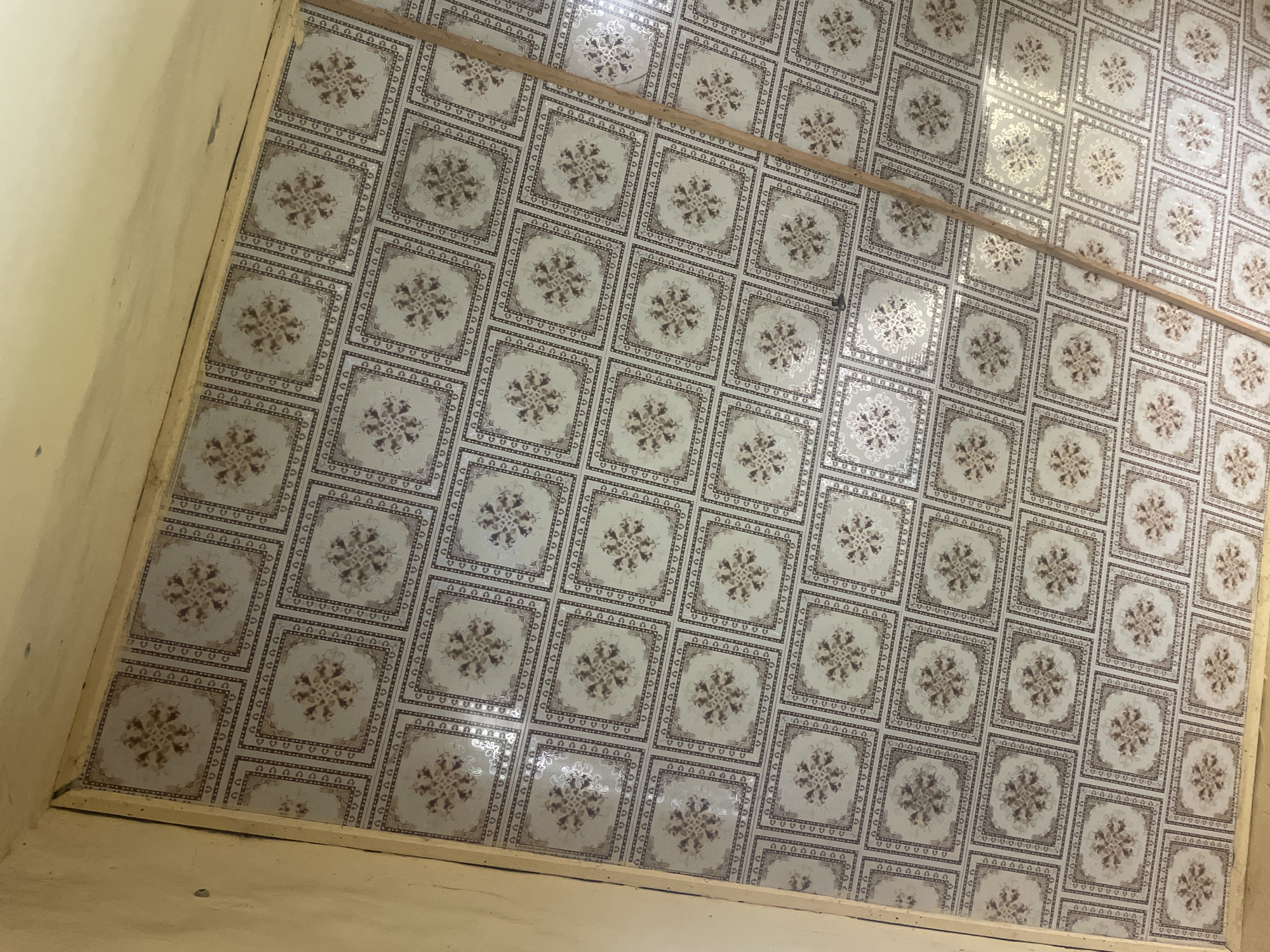 The image shows a close-up, overhead view of a tiled floor in a room. The tiles are square and have a repeating pattern of a central floral motif surrounded by decorative borders. The color palette of the tiles is predominantly light gray and beige, with darker brown accents in the borders and the floral design. The tiling appears to be in good condition, with a consistent pattern across the visible area.
A thin, light-colored wooden plank runs diagonally across the center of the image, dividing the tiled surface. The plank appears to be a decorative element or possibly part of a shelf or divider. The walls of the room are visible on the left and bottom edges of the image. The wall on the left is painted a light yellow or cream color and shows some minor scuff marks or dirt. The wall at the bottom appears to be the base of the room, possibly a skirting board or the transition to another surface.
The lighting in the image suggests an indoor setting, possibly with natural light coming from an unseen source, as there are some subtle reflections on the tiled surface. There is no indication of people or any specific activity taking place. The overall impression is that of a simple, functional interior space. The image is taken from a high angle, looking down onto the floor. The context provided indicates the location is Jalingo, Nigeria, but there are no specific visual cues in the image to confirm this detail. No text is visible in the image.