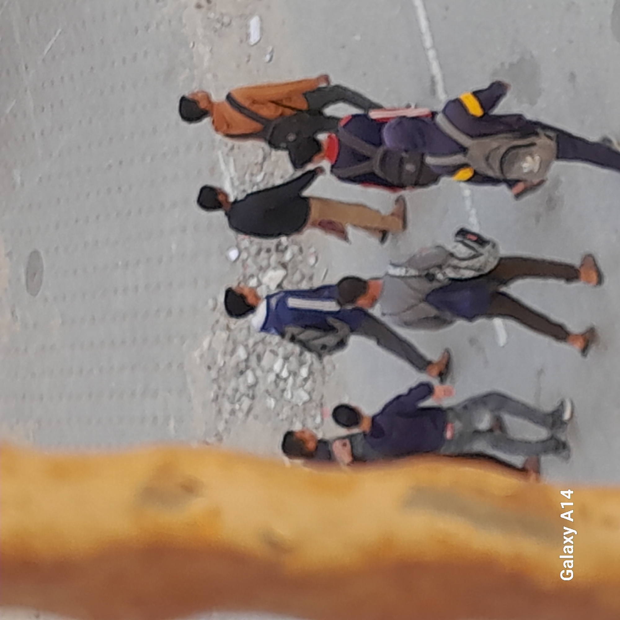 An elevated, downward-angled photograph captures a group of approximately seven individuals, appearing to be adult males, moving or standing on a paved surface. The ground is predominantly grey, marked by faint white lines and patches of scattered lighter-colored debris, resembling small stones or gravel.
The individuals are dressed in casual attire, including various colored shirts or jackets (orange, black, blue, grey) and trousers (tan, black, grey). Several are carrying backpacks, one distinctly blue and grey with yellow accents, and another grey and white. Their heads, mostly dark-colored, suggest either dark hair or head coverings. The group generally faces towards the left side of the frame.
The image was taken during daytime, with no specific weather conditions evident. The immediate foreground is dominated by a large, blurry, yellowish-brown object, which partially obscures the lower parts of the subjects and suggests the photograph was taken from behind a railing or similar structure. Due to the distance and resolution, individual emotions are not discernible. The text "Galaxy A14" is visible vertically in the bottom right corner of the image.