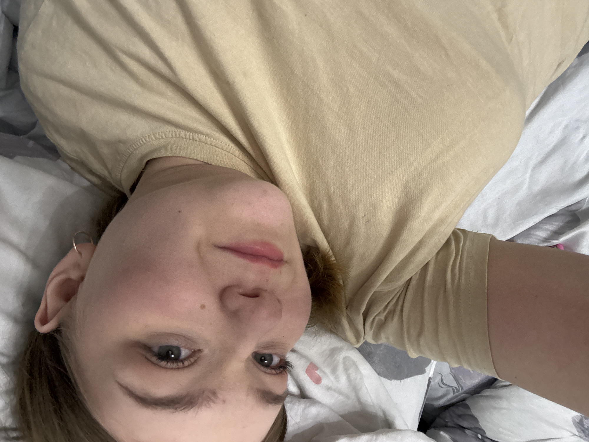 A person is lying on a bed. The person has pale skin, light brown hair, and grey eyes. They are wearing a tan t-shirt. The bed is covered in white sheets with grey and floral patterns. The location is not specified.