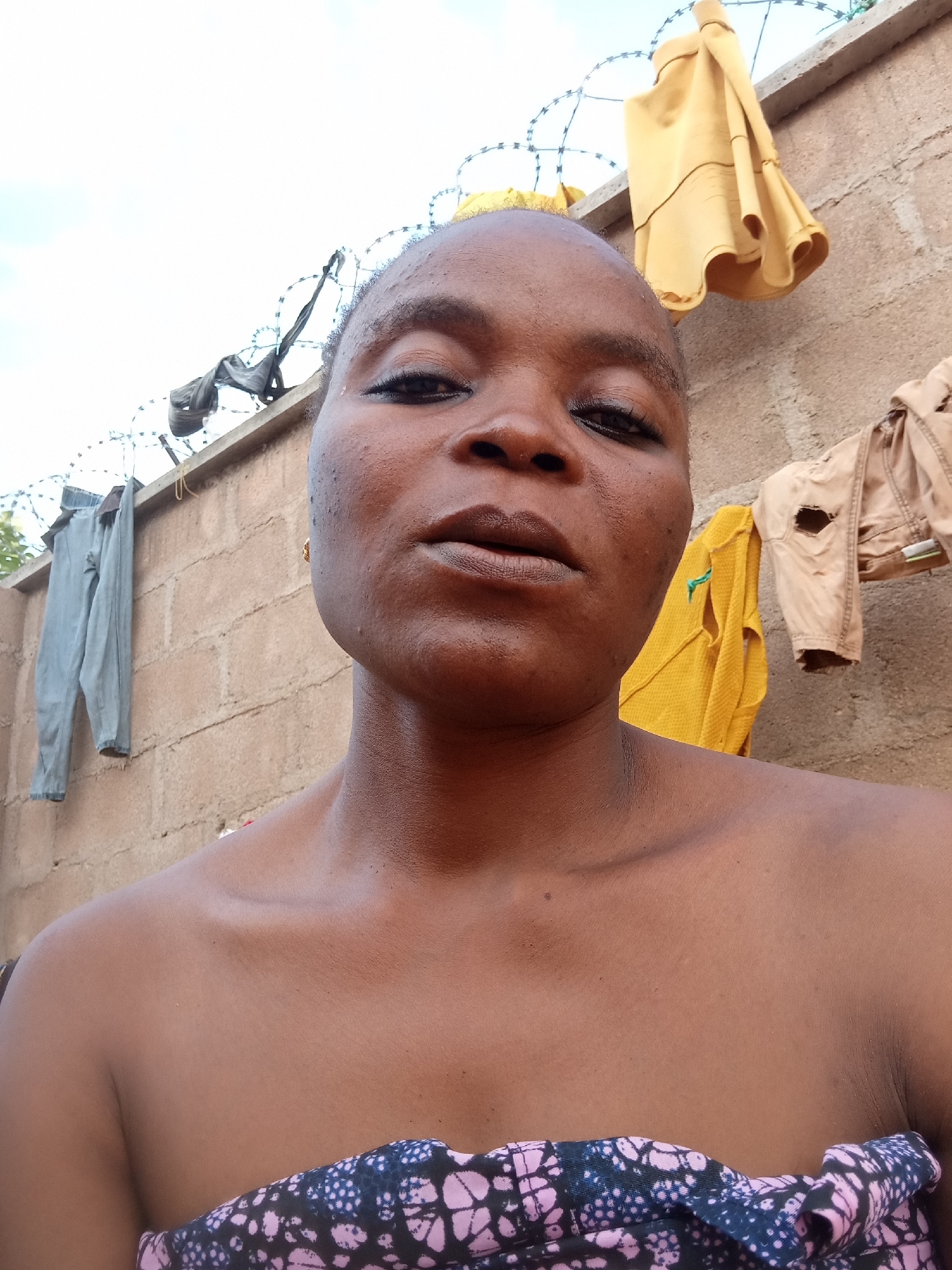 A close-up, selfie-style photograph shows a Black woman with a shaved head looking directly at the camera. She is wearing a patterned fabric wrapped around her chest. Behind her, a brick wall is visible, with barbed wire strung along the top. Several items of clothing are hanging on the wall: a pair of grey pants, a yellow garment, and a tan garment with a hole. The background consists of a pale sky with some clouds. The overall setting appears to be an outdoor area, possibly a residential exterior in Kason, Nigeria, given the presence of laundry hanging to dry and the architectural style of the wall.
