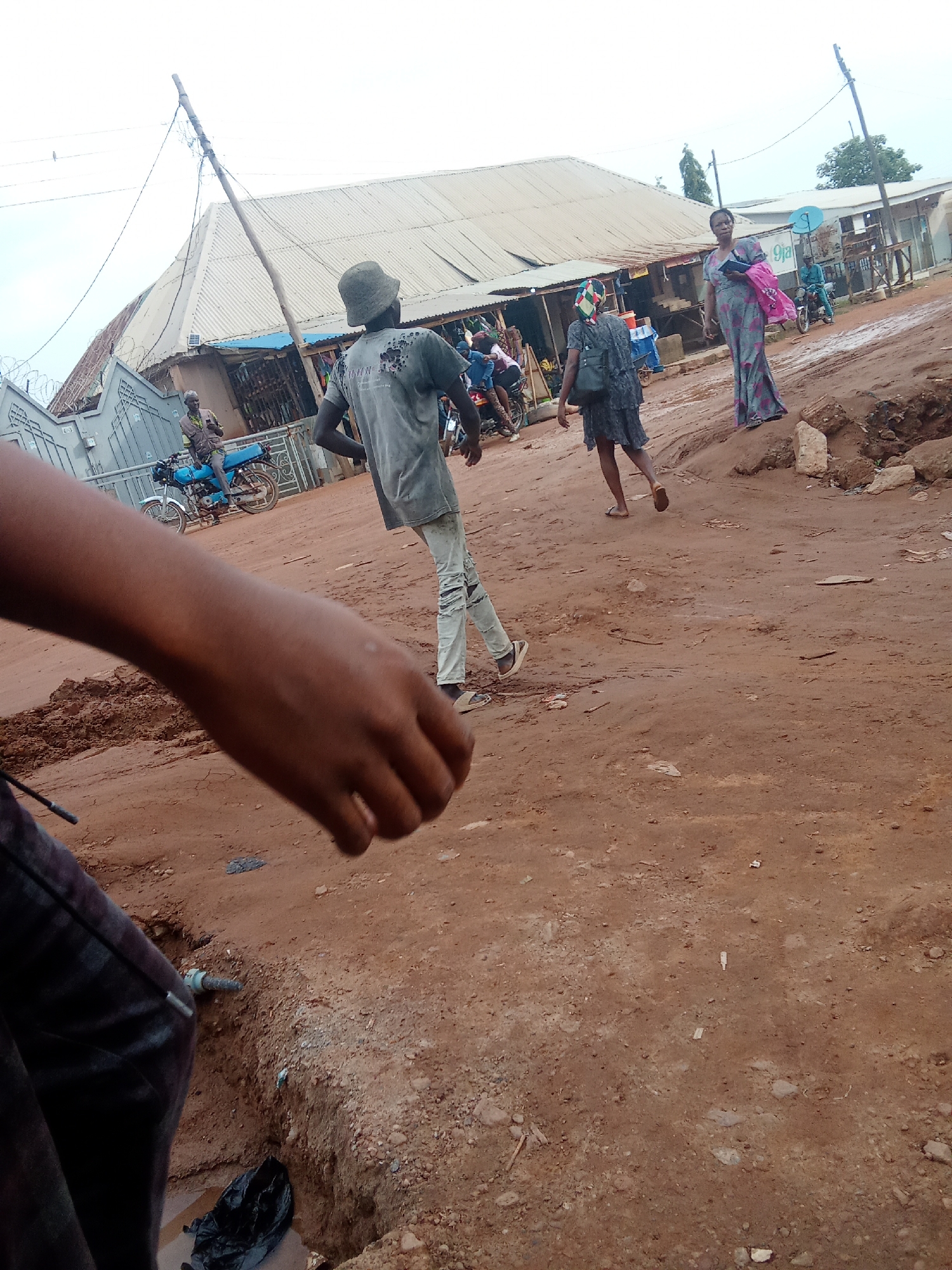 The image depicts an outdoor scene in a dusty, urban environment. In the foreground, a person's arm and part of their torso are visible, wearing dark patterned pants. The ground is primarily composed of reddish-brown dirt with scattered stones and debris, including a black plastic bag and a section of exposed pipe.
Further into the scene, a young man wearing a bucket hat and a grey t-shirt with a design on the back walks away from the camera. He is wearing ripped light-colored pants and sandals. To his right, a young person wearing a dark dress and a backpack walks away from the camera, also in sandals.
In the background, a woman in a long, patterned dress and carrying a pink bag walks towards the right side of the image. Behind her, a man on a blue motorcycle is visible.
Several buildings with corrugated metal roofs line the street. There are also utility poles with wires. The overall atmosphere appears to be that of a busy street in a developing area, possibly a market. The sky is overcast. The specific location is identified as Unknown, Unknown.