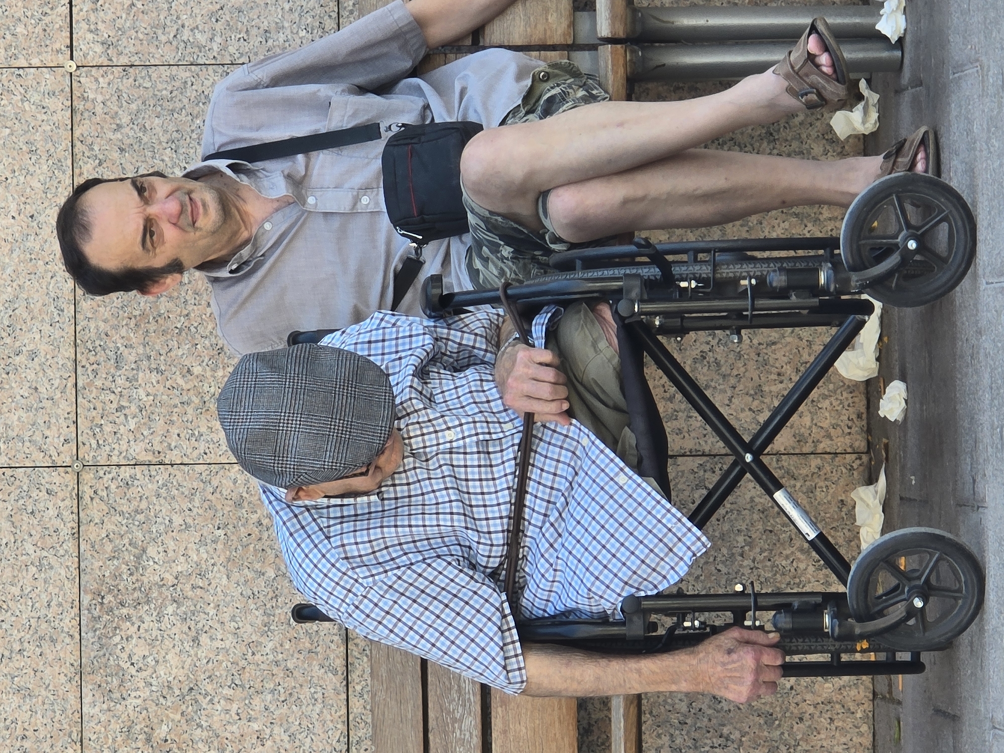 Two men are seated on a wooden bench in front of a granite wall. The man on the left is an elderly individual wearing a plaid shirt and a flat cap, seated in a wheelchair. He is leaning forward, with his head down and his right hand resting on the armrest of the wheelchair. The man on the right is younger, wearing a light-colored button-down shirt and camouflage shorts. He is looking to his left with a neutral expression. He has a small black bag slung across his body. Both men's legs are visible, and the younger man's legs are crossed. The ground in front of the bench has scattered white paper or tissue. This scene appears to be outdoors, likely in a public area, possibly Cornellà De Llobregat, Spain, given the information provided.