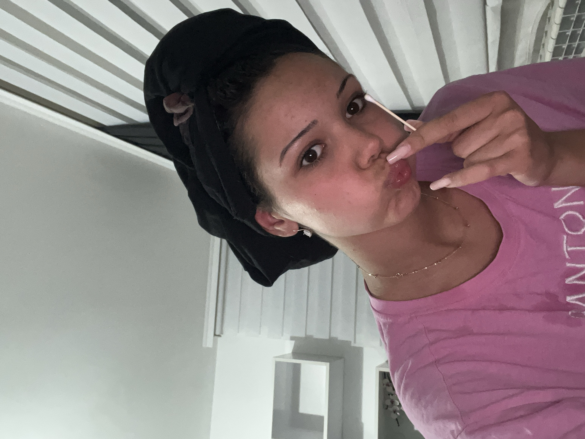 The image captures a close-up portrait of a young female subject indoors. She appears to be in her late teens or early twenties with light-to-medium skin, dark brown eyes, and dark hair pulled back and covered by a black headwrap or bonnet. A small section of a light pink or faded fabric is visible at the top of the headwrap. She is wearing a pink short-sleeved t-shirt. A delicate gold chain necklace with small, circular elements adorns her neck, and a single drop earring featuring a clear stone hangs from her right ear. Her fingernails are long and painted in a light pink or nude shade.
The subject is holding a white cotton swab (Q-tip) between her right thumb and index finger, with the tip touching her pursed, glossy lips, creating a pouting expression. She is looking directly at the camera.
The background features a light-colored, possibly white or light grey, wall. To the left, two white, rectangular, open shelves are mounted on the wall, appearing largely empty. To the right and behind the subject, a window covered by horizontal white slatted blinds (venetian blinds) is visible, with the slats partially open. The lighting is even and consistent with an indoor environment. There are no visual elements in the image to indicate a specific geographical location such as Zlatograd, Bulgaria.