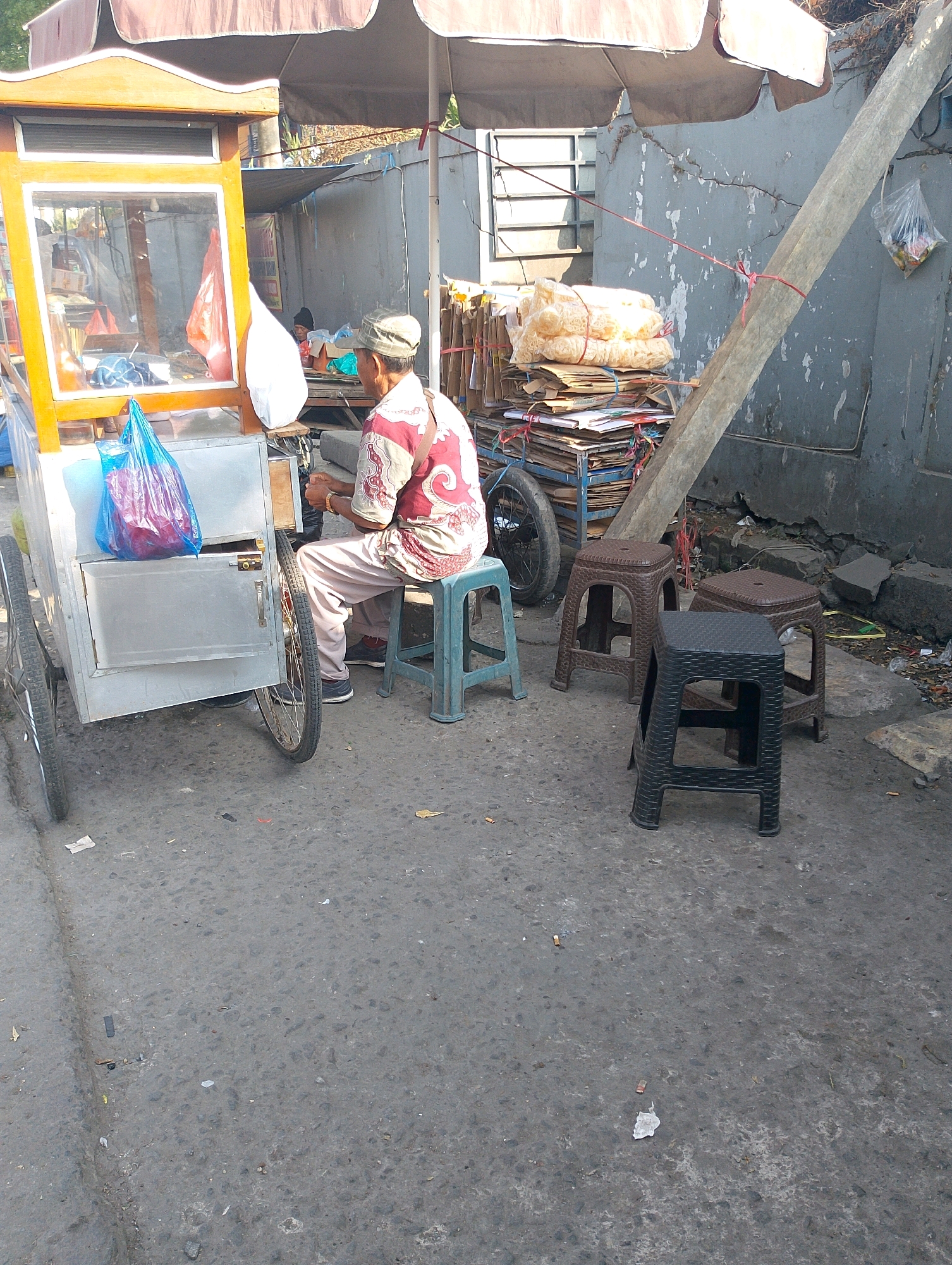 A man, wearing a cap and a patterned shirt, sits on a green plastic stool next to a food cart. The cart, made of metal and wood, has a display window and is parked on an asphalt road. Behind the cart, stacks of cardboard and packaged food items are piled high, secured with red string. Several plastic stools, two brown and one black, are arranged in front of the food cart. A large, slanted wooden pole stands to the right of the scene, and a grey concrete wall forms the backdrop. In the distance, foliage and a window with metal bars are visible. The environment appears to be an outdoor urban setting, possibly in Lembang, Indonesia, given the context. The ground is littered with debris.