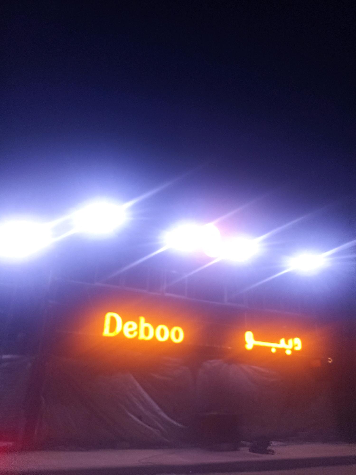 An outdoor, nighttime photograph captures a neon sign that reads "Deboo" in English and what appears to be Arabic script on the right side. The sign is illuminated in bright yellow-orange light. Above the sign, several powerful spotlights cast intense white light, creating lens flares and washing out parts of the background. The overall scene suggests an event or establishment at night. The background is dark blue, indicating the night sky. In the lower portion of the image, a dark, possibly fabric-covered structure is visible beneath the neon sign, along with a dark-colored ground or pavement. The photograph is likely taken in Gaza, Palestinian Territories, due to the presence of Arabic script and the context of such businesses in the region.