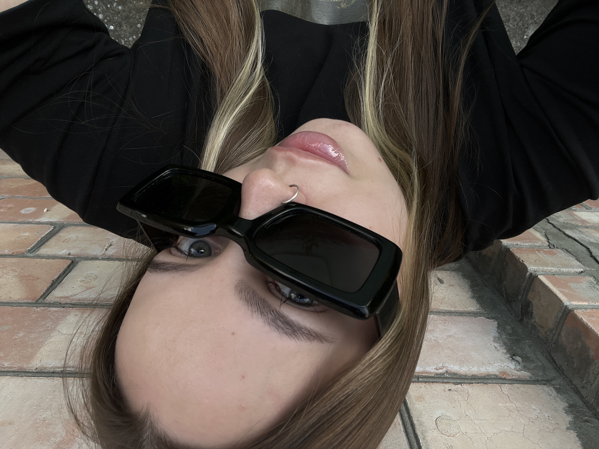 A young woman with long, light brown hair, partially dyed blonde, wears black sunglasses and a black long-sleeved shirt. She has a silver nose ring. She is positioned close to a brick wall, with her face tilted upwards and towards the camera. Her lips are slightly parted and glossy. The background consists of the textured brick wall and a darker, rougher surface below, possibly concrete or asphalt.
