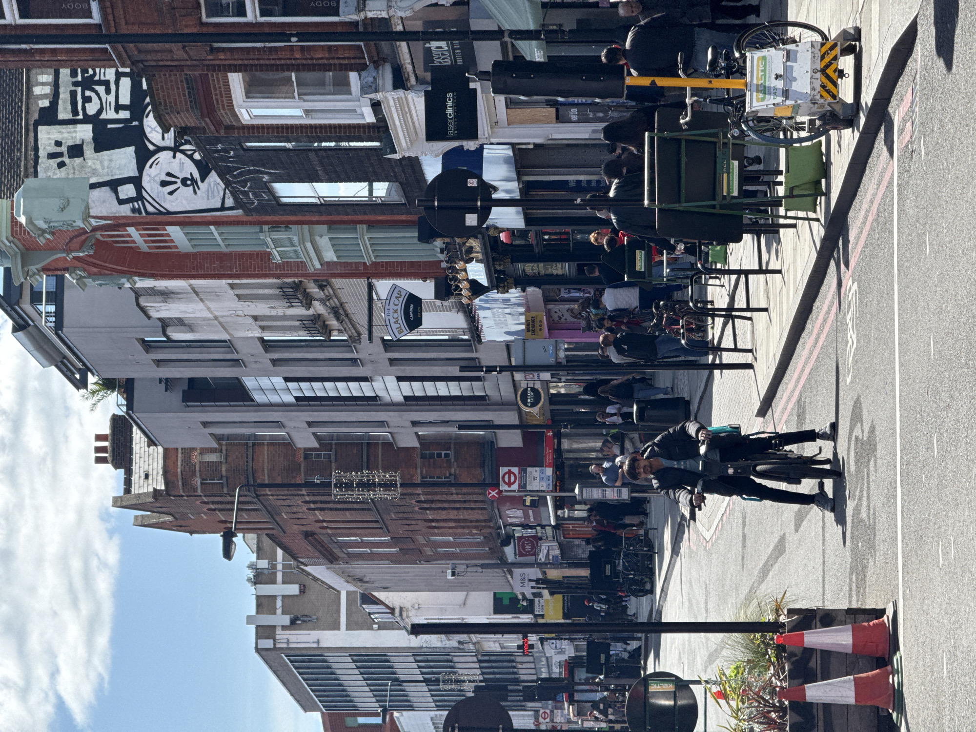 This image depicts a street scene in London, United Kingdom. The buildings on either side of the street are multi-storied, with a mix of architectural styles including brick and modern glass facades. The sky is blue with some clouds visible. Several people are walking on the sidewalks, and some are sitting at outdoor cafe tables. A person is standing with a bicycle. Street signs, traffic lights, and commercial signs are visible, including "M&S" (Marks & Spencer), "Pret a Manger", "THE BLACK CAP CAMDEN" and "laserclinics". Traffic cones are present at the edge of the road.