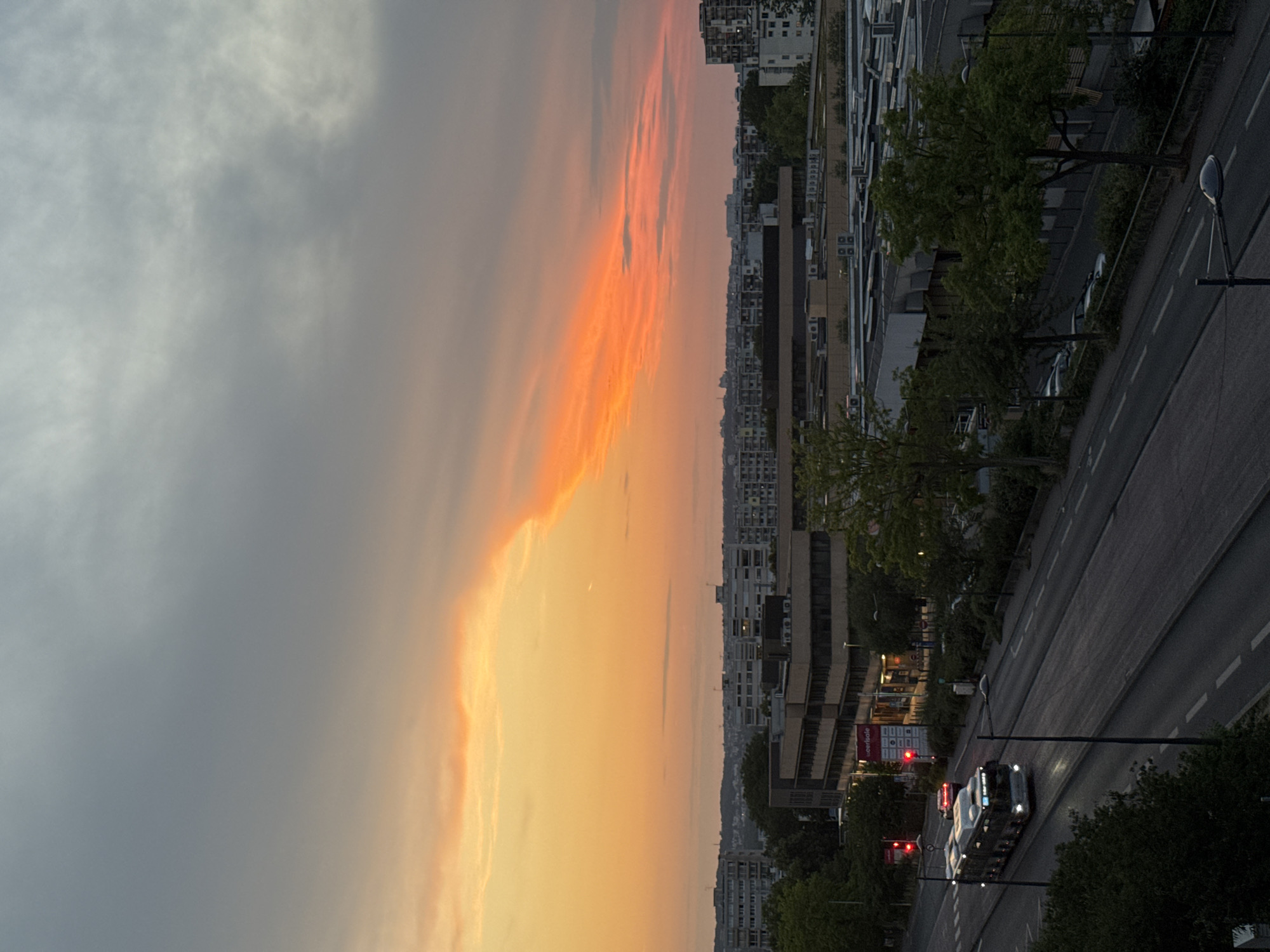 An aerial medium shot captures a street scene in an urban environment during a sunset. A long, articulated bus with its headlights on travels down the left lane of a multi-lane road. A red sports car is visible behind the bus. Traffic lights are illuminated red. On the right side of the road, trees line a sidewalk. In the background, multi-story buildings and apartments form a city skyline. The sky above displays a dramatic sunset with streaks of orange and yellow clouds against a backdrop of gray clouds. The lighting suggests either dusk or dawn.