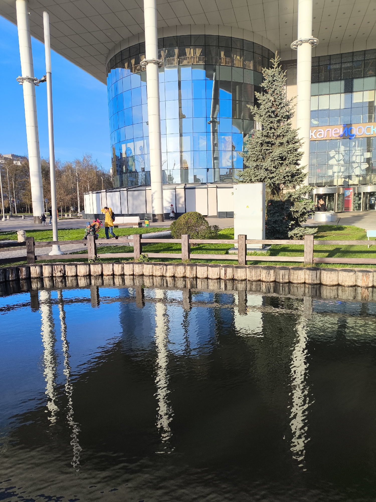 This image depicts a vibrant urban scene in Moscow, Russia, bathed in bright daylight under a clear blue sky. A prominent modern building with a curved, reflective blue glass facade and large, white cylindrical support pillars dominates the background. The building's glass reflects the bright sky and elements from the surrounding environment. On its facade, the Cyrillic text "Калейдоскоп" (Kaleidoscope) is visible, indicating a commercial establishment like a shopping center.
In front of the building, a well-kept grassy area features a small evergreen tree and some bushes. A white utility box is situated near the tree. Several people can be seen on a paved walkway further back, including an individual wearing a distinct bright yellow jacket, walking with another person.
The foreground is occupied by a calm body of water, which provides a striking reflection of the building's pillars and the blue sky, creating shimmering, elongated patterns. A rustic wooden fence, constructed from horizontal planks and vertical posts, borders the grassy bank, separating it from the water. Along the immediate edge of the water, a barrier fashioned from vertically arranged logs is visible. The strong reflections in both the water and the building's glass are a notable detail, enhancing the sense of light and space on this sunny day.