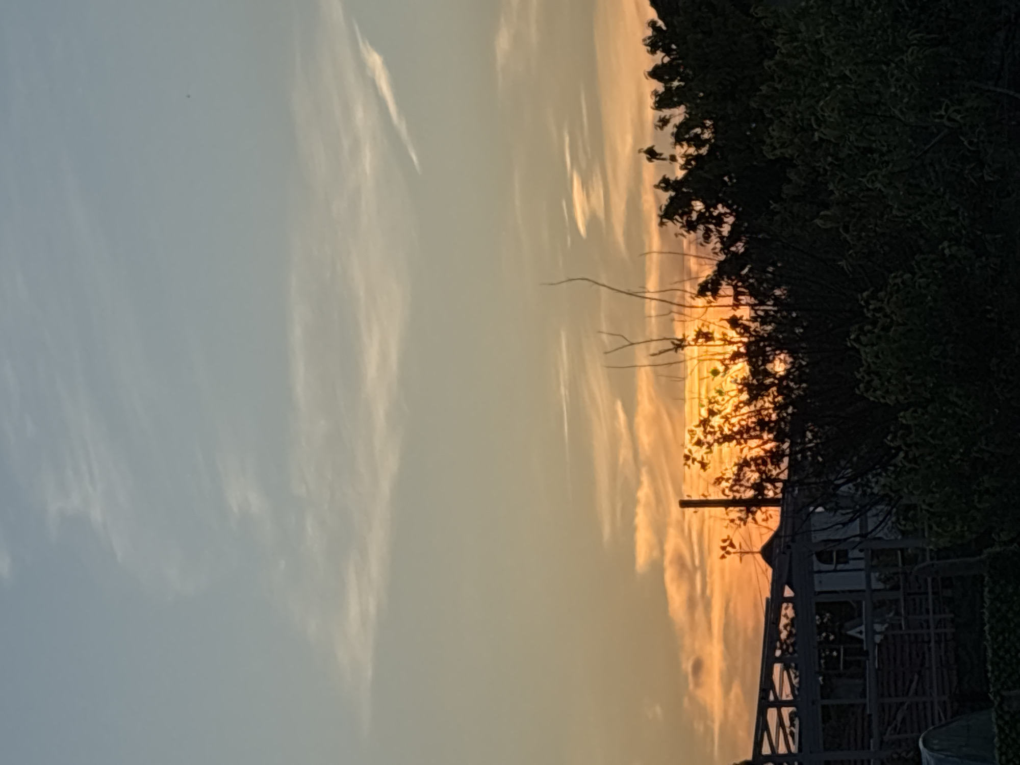 The image depicts a sunset in Unknown, Unknown. The sky is a gradient of colors, transitioning from blue on the left to orange on the right. Wispy clouds are visible in the sky. A tree with dense foliage is on the right, partially obscuring the sunset. In the lower right, there is a structure, possibly a building or framework, with geometric lines and shapes. The light of the sunset shines through the branches and the structure, creating a play of light and shadow.