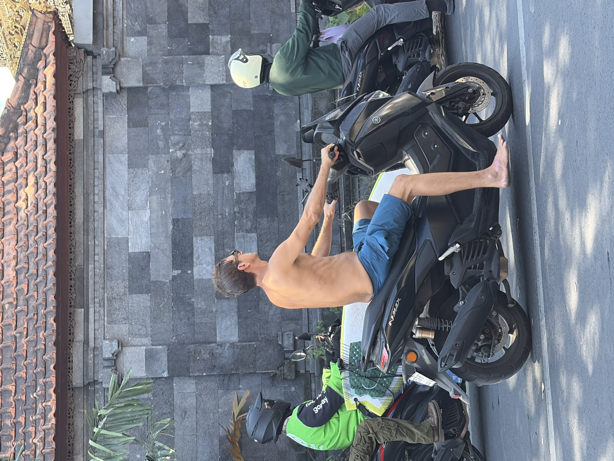 A shirtless man wearing blue shorts rides a black Yamaha NMAX scooter. He has a surfboard strapped to the back of his scooter. Beside him, a person wearing a green jacket and black helmet on a scooter carries a surfboard. Further down the road, a person in a green long-sleeved shirt and cream helmet is on a scooter. The scene appears to be on a street in North Kuta, Indonesia, with a grey stone wall and tiled roof in the background.