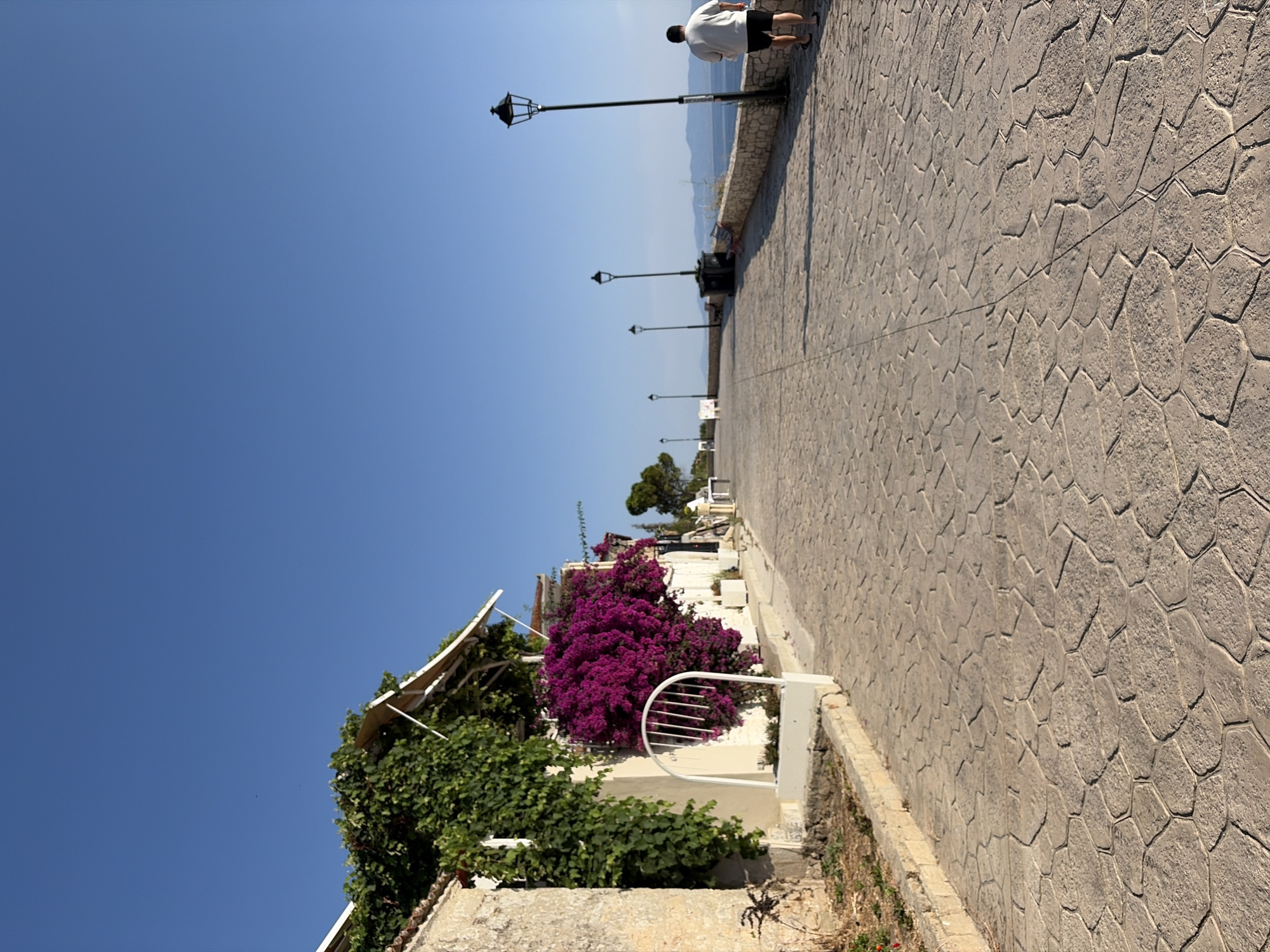 A person, appearing to be a male, walks away from the camera on a wide, stone-paved promenade. He is wearing a white t-shirt and dark shorts. The promenade runs alongside a low stone wall, beyond which water and distant landmasses are visible under a clear blue sky. The promenade is lined with several ornate, black lampposts. On the left side of the promenade, a building is visible, adorned with vibrant purple bougainvillea flowers and green vines. The building features a white awning. The paving stones on the promenade have a hexagonal pattern.