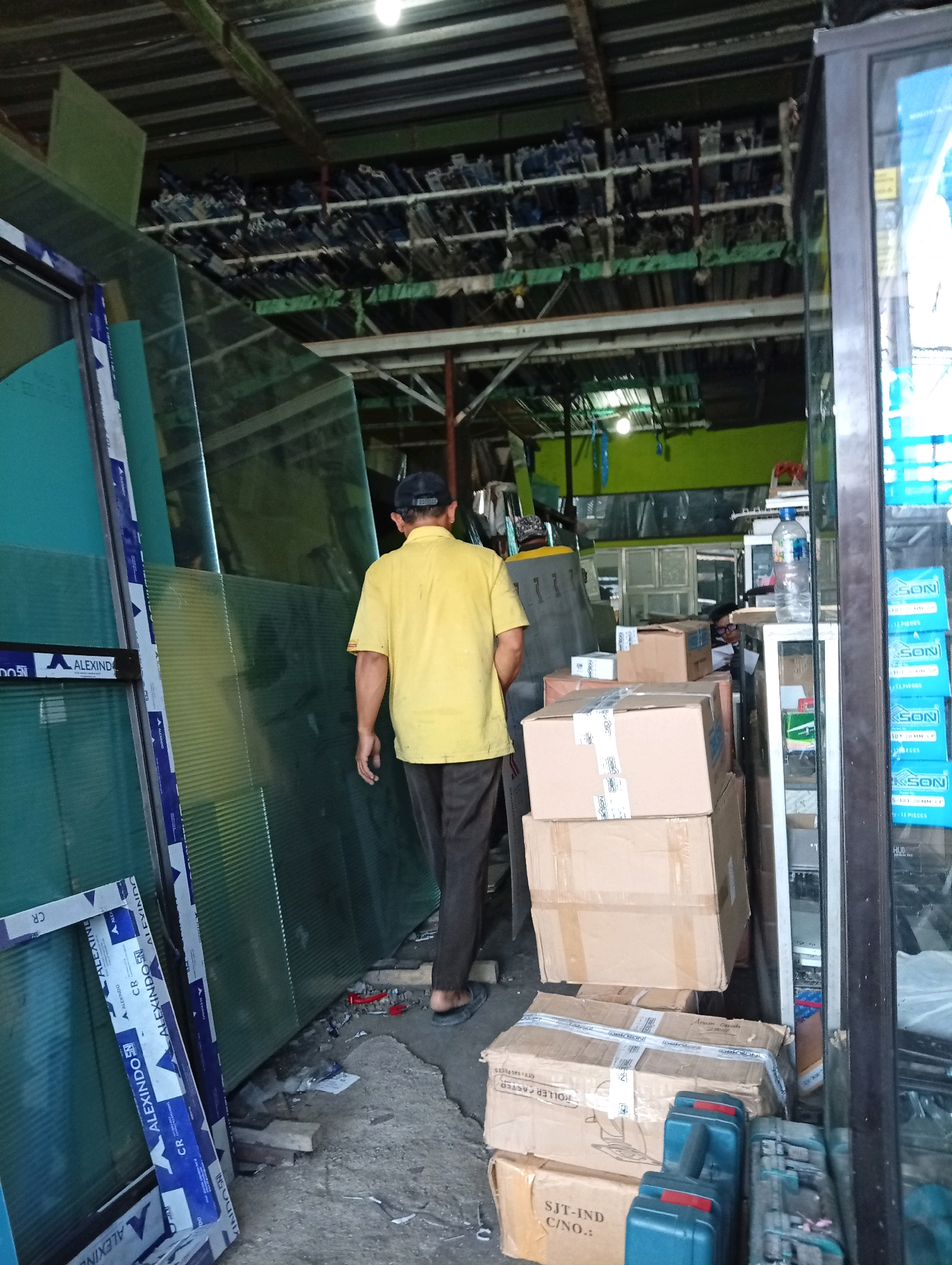 In the city of Bangah, Indonesia, a person wearing a yellow t-shirt and a dark baseball cap walks away from the camera. The individual is in a warehouse-like setting, with large sheets of what appear to be green plastic or glass stacked vertically on the left side of the frame. The person is walking between these sheets and a wall of shelves filled with various items in the background. Several cardboard boxes are stacked on the floor in the mid-ground. A glass-fronted display case or refrigerator is visible on the far right, with blue packaging for a product labeled "JKSON" visible inside. The ceiling appears to be made of corrugated metal, and the lighting is dim, suggesting an indoor storage or retail space.