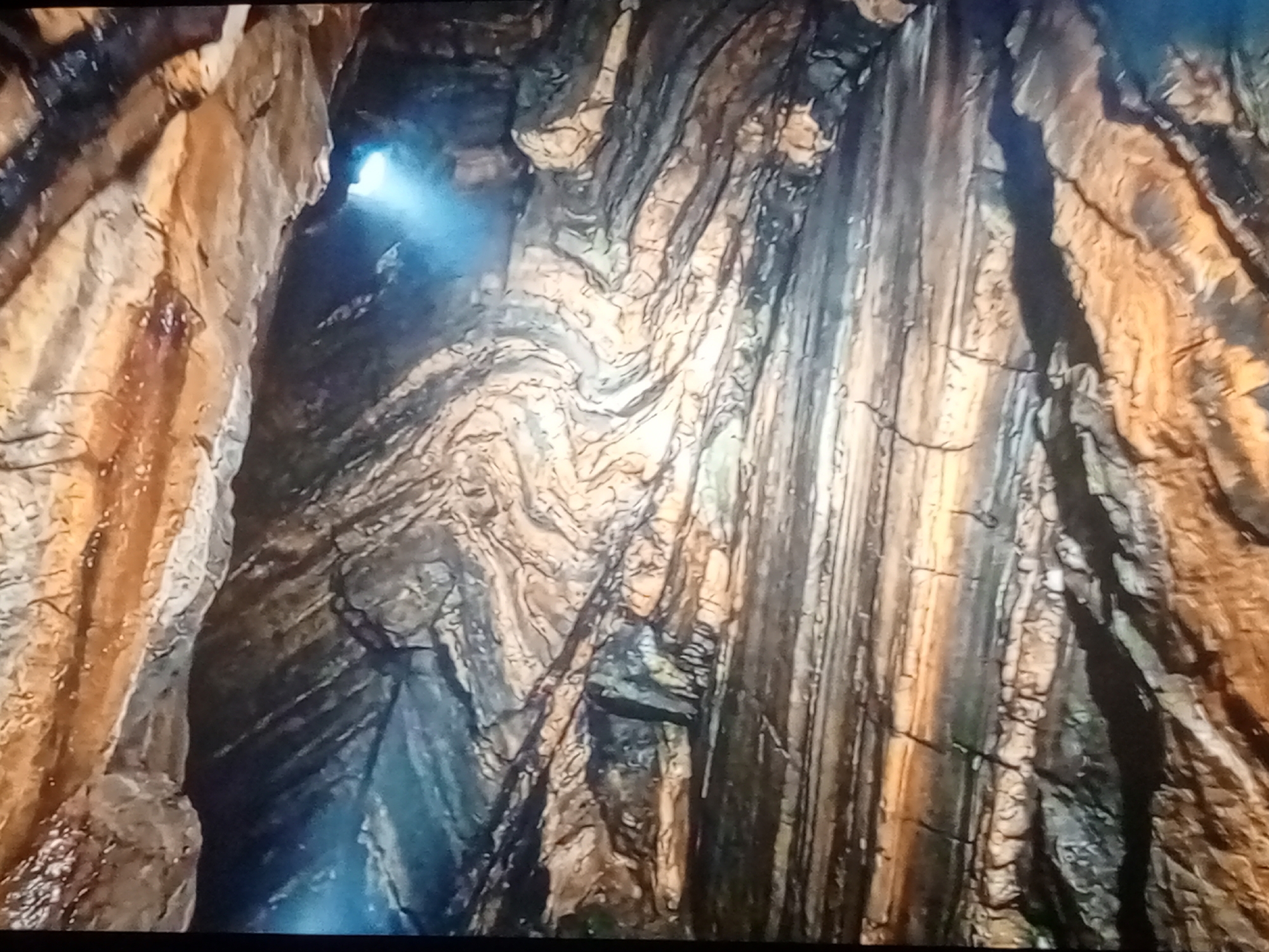 The image shows a dimly lit cave or underground natural formation with intricate rock layers. Light emanates from an opening at the top left, casting a blue glow on the wet, textured surfaces of the rock. The rock walls are folded and striated, displaying a variety of earthy tones including browns, grays, and whites. The overall impression is one of a geological wonder, possibly a cave system or a natural fissure.