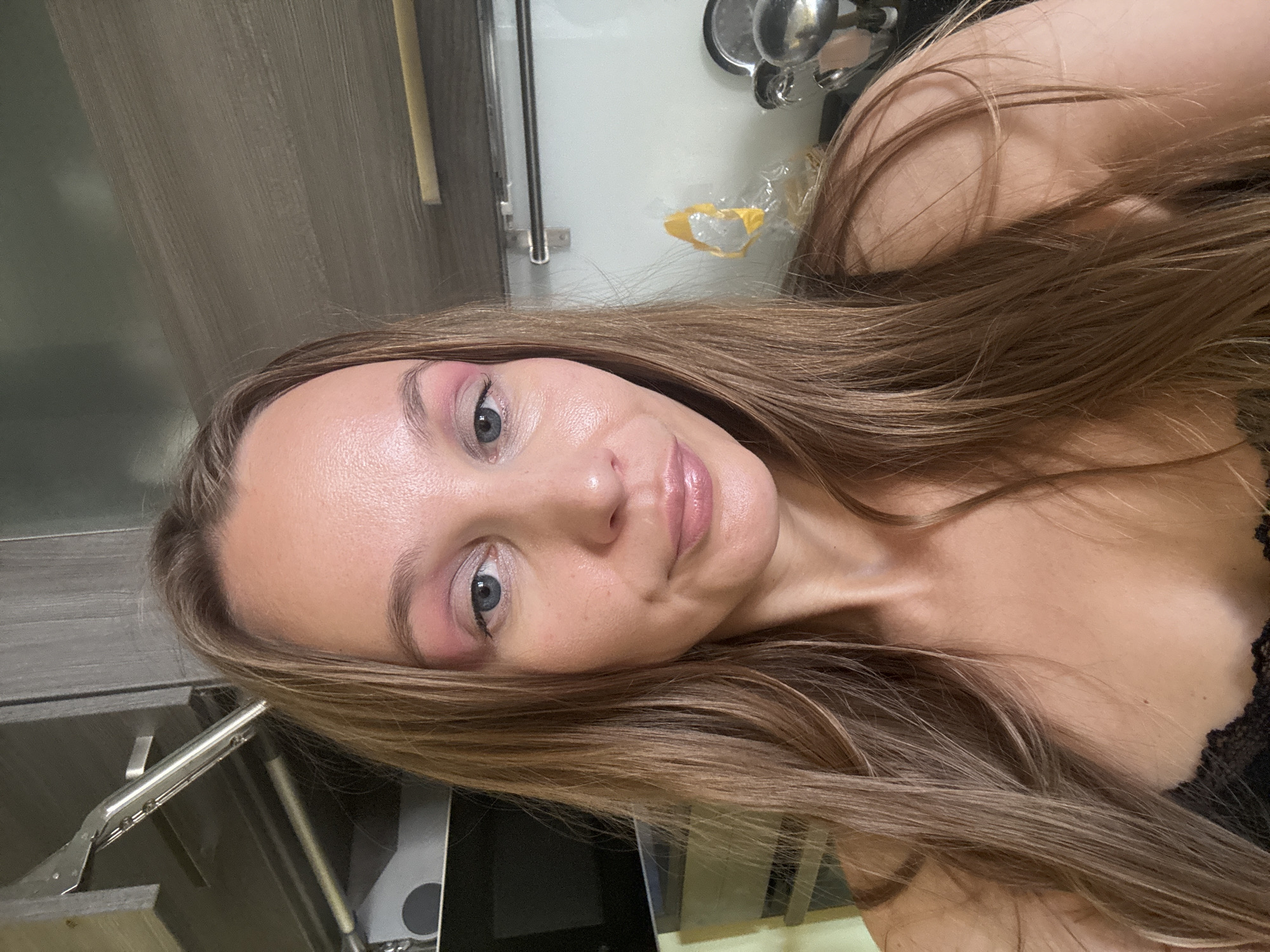 A young woman with long, blonde hair and blue eyes is pictured in a selfie. She is wearing pink eyeshadow and lip gloss. She is in a kitchen, with cabinets and appliances visible in the background. The overall setting appears to be a home. The image does not contain any specific details that would allow for the identification of the location within the city of Überherrn, Germany.