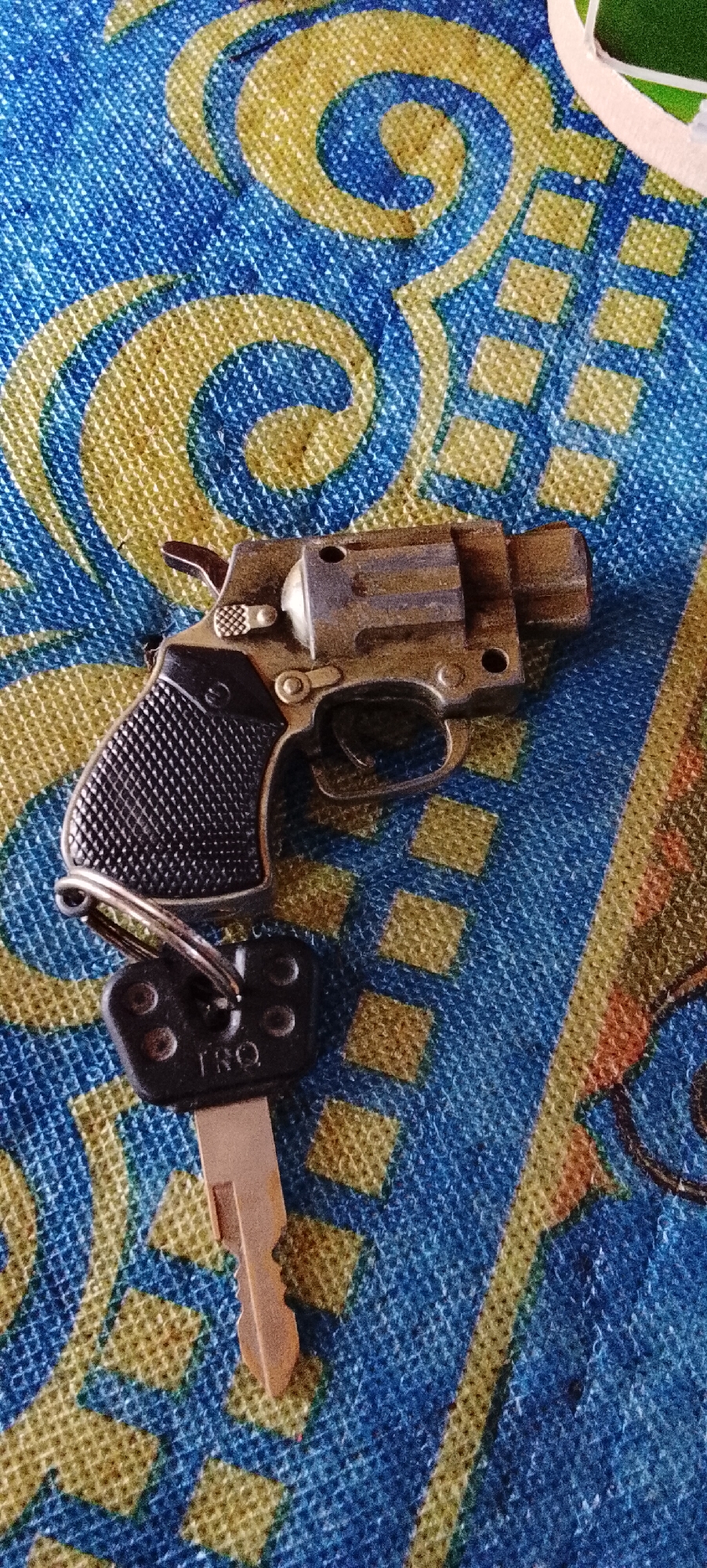 The image displays a close-up, high-angle shot of a keychain that includes a miniature replica of a revolver and a car key, resting on a blue and yellow patterned fabric. The fabric has a textured appearance with swirling yellow designs and a grid of yellow squares on a blue background. The toy gun is metallic with a black grip and a silver hammer. The car key is made of metal with a black plastic head that has four circular indentations and the letters "TRQ" embossed on it. The image does not provide any information or context related to the city of Padang Dalom, Indonesia.