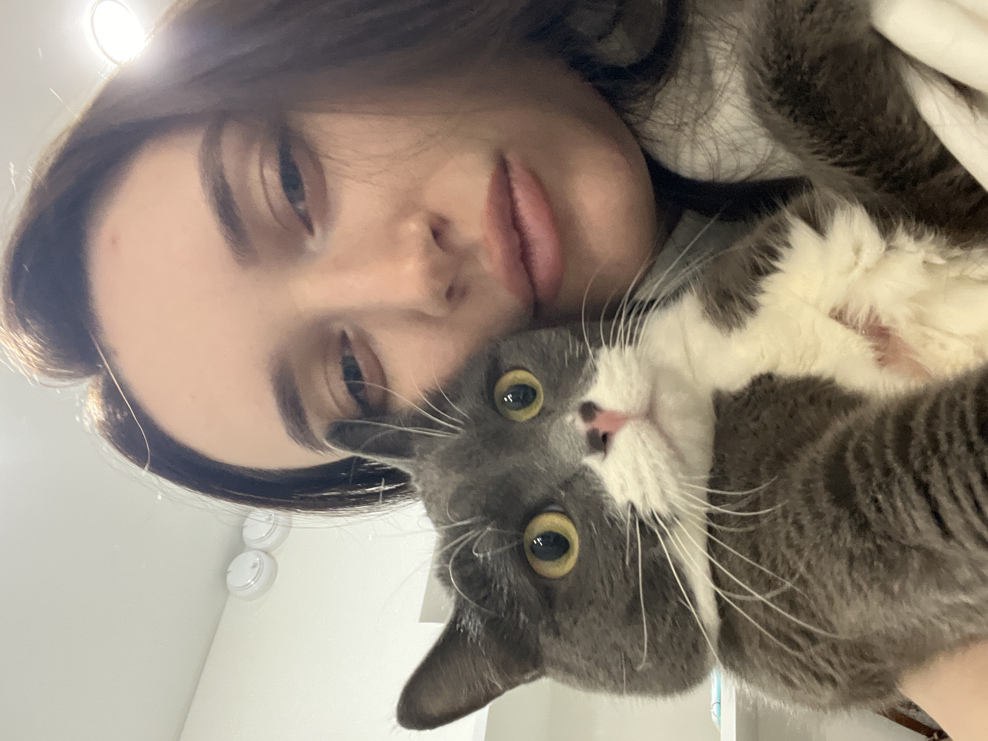A selfie shows a young woman holding a grey and white cat. The woman has dark brown hair, fair skin, and pink lips. Her eyes are half-closed. She is wearing a light-colored top. The cat has yellow eyes and a pink nose. The background appears to be a white or light-colored wall. No specific details indicate that the location is Tyumen, Russia. The environment is indoors. The interaction is between a person and a pet.
