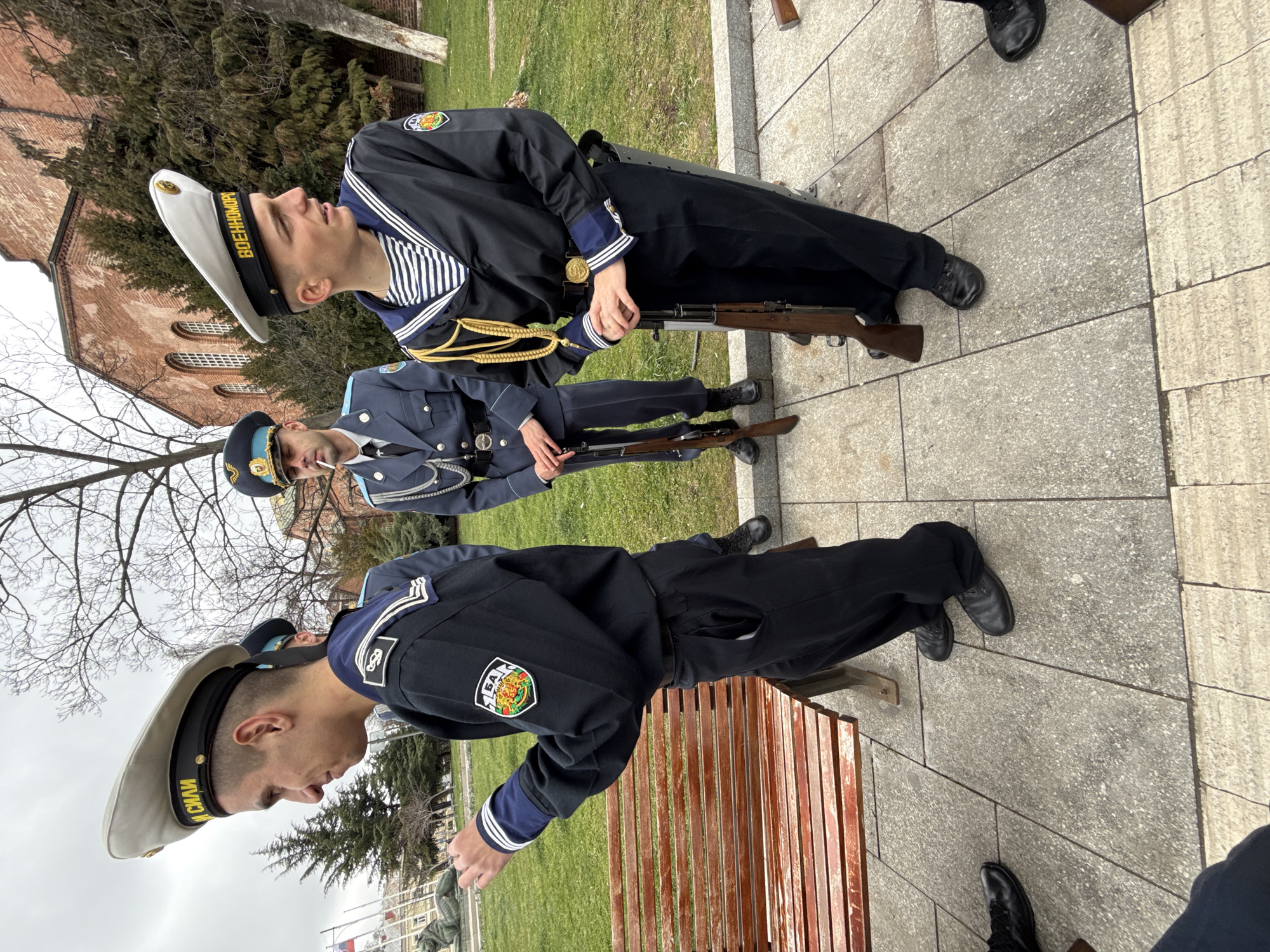 The image shows three military personnel standing in a park, two in navy uniforms with white sailor hats and striped shirts, and one in a light blue military uniform with a peaked cap. Each holds a rifle. The setting appears to be in Sofia, Bulgaria, recognizable by the brick building behind them, reminiscent of the architecture of the St. Alexander Nevsky Cathedral. They are standing on a stone-paved area next to a patch of grass. One is leaning on a wooden bench.