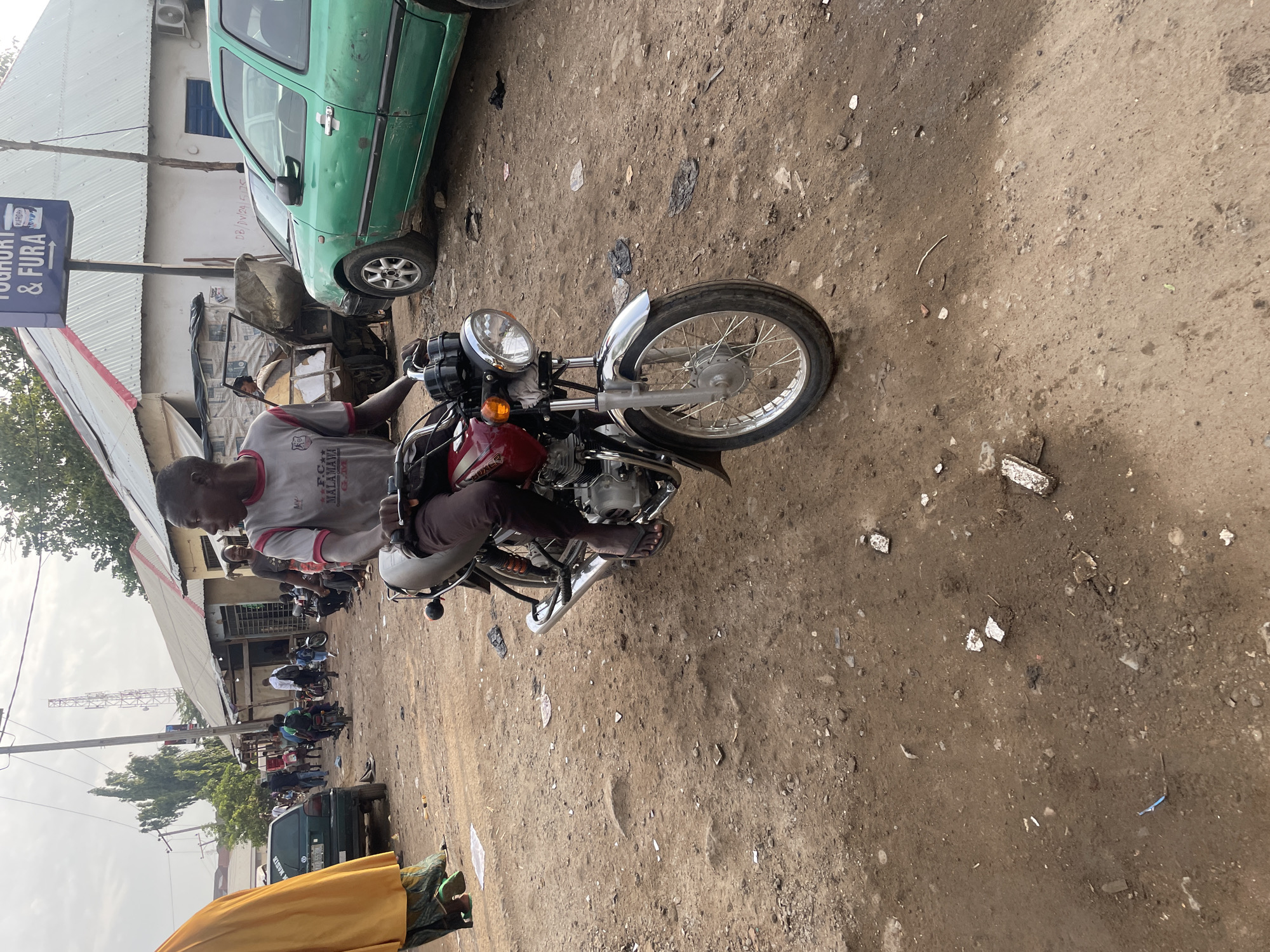 The image depicts a man sitting on a red motorcycle parked on a dirt road in a commercial area of Abuja, Nigeria. The man is wearing a gray t-shirt, brown pants, and sandals. He appears to be looking at his phone. The motorcycle is parked near a building with a sign that reads "Yoghurt & Fura". A green car is parked nearby. Other people and motorcycles are visible in the background, along with various buildings and a cell tower. The road is unpaved with debris scattered on the ground.