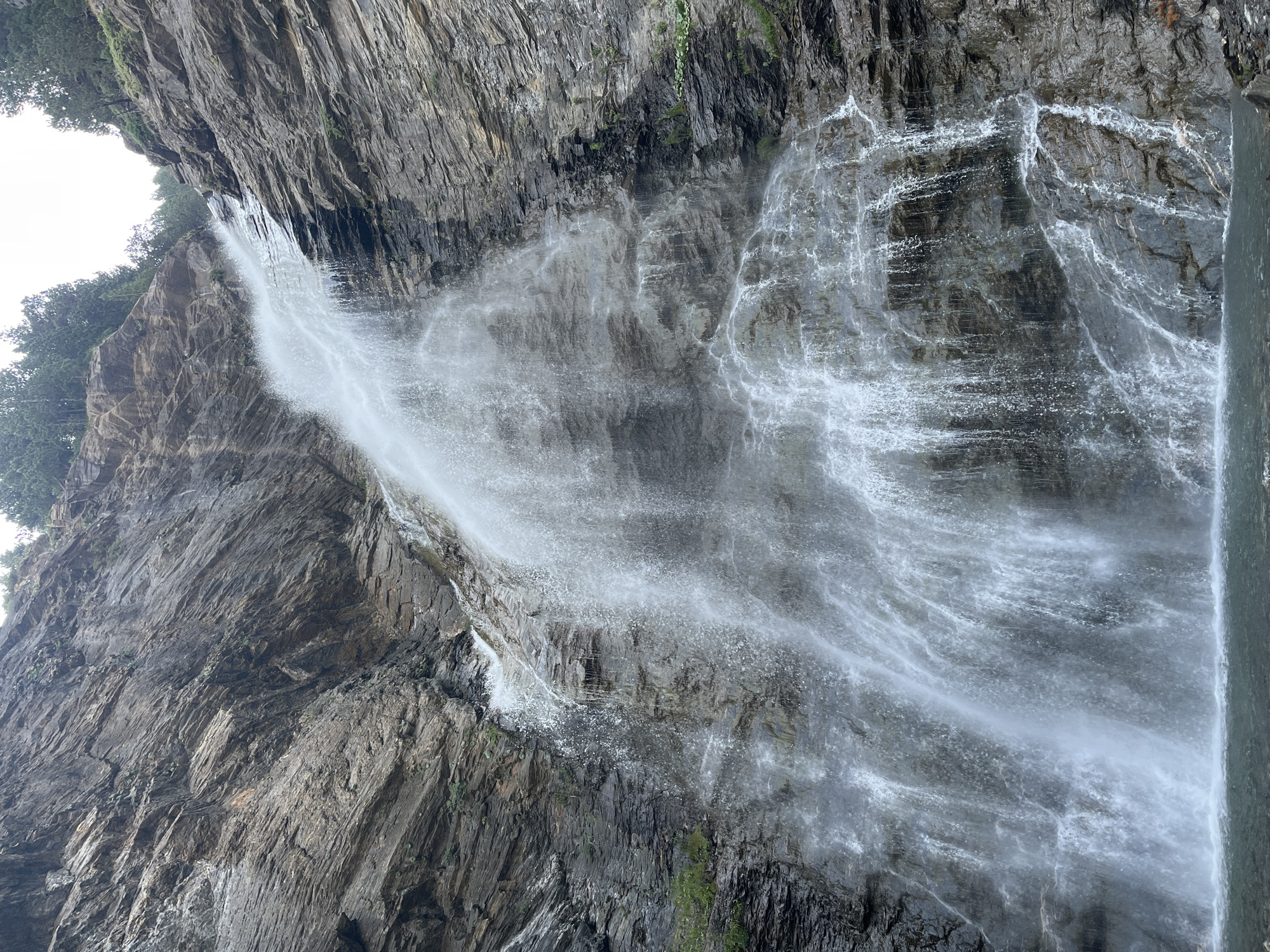 The image shows a tall, cascading waterfall in a rocky gorge. The water is white and foamy as it plunges down the steep, gray rock face. The rock is layered and appears rugged, with some patches of green vegetation clinging to the sides. At the top of the gorge, a line of dark green evergreen trees is visible against a bright, overcast sky. The bottom of the waterfall spills into a pool of calm, dark water. This appears to be the Congosto de Ventamillo, located in the vicinity of Benasque, Spain.