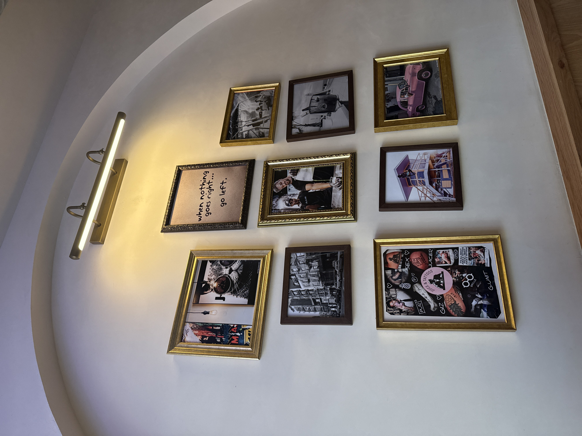This indoor shot from Mexico City, Mexico, captures a brightly lit wall adorned with nine framed pictures of varying sizes and orientations, creating a gallery display. A long, horizontal, gold-toned light fixture is mounted on the left, casting a warm, bright glow across the light-colored wall and the top left portion of the picture arrangement. An arched architectural detail is visible in the upper left background.
The framed art includes a mix of black and white and color photographs, as well as text-based art. Notable subjects in the pictures include: a black and white image of a pink vintage car seen from above; a black and white image of two smiling individuals; a person diving from a colorful structure into water; a detailed black and white street scene with buildings; and a close-up of a cat wearing sunglasses. One frame features the motivational quote, "when nothing goes right... go left." Another frame on the bottom right contains a dense collage of colorful stickers and text, including phrases like "TATTOOS," "JUICE & TATTOOS" (repeated), "ITS ALL ABOUT JUICE & TATTOOS" (repeated), "G-UNIT," "SEX," "PUR," "GIZ," "BRILLE," and "JUICER." Some frames are golden or brass-colored, while others are dark brown. The overall scene suggests a contemporary and curated aesthetic, possibly within a cafe, bar, or residential setting.