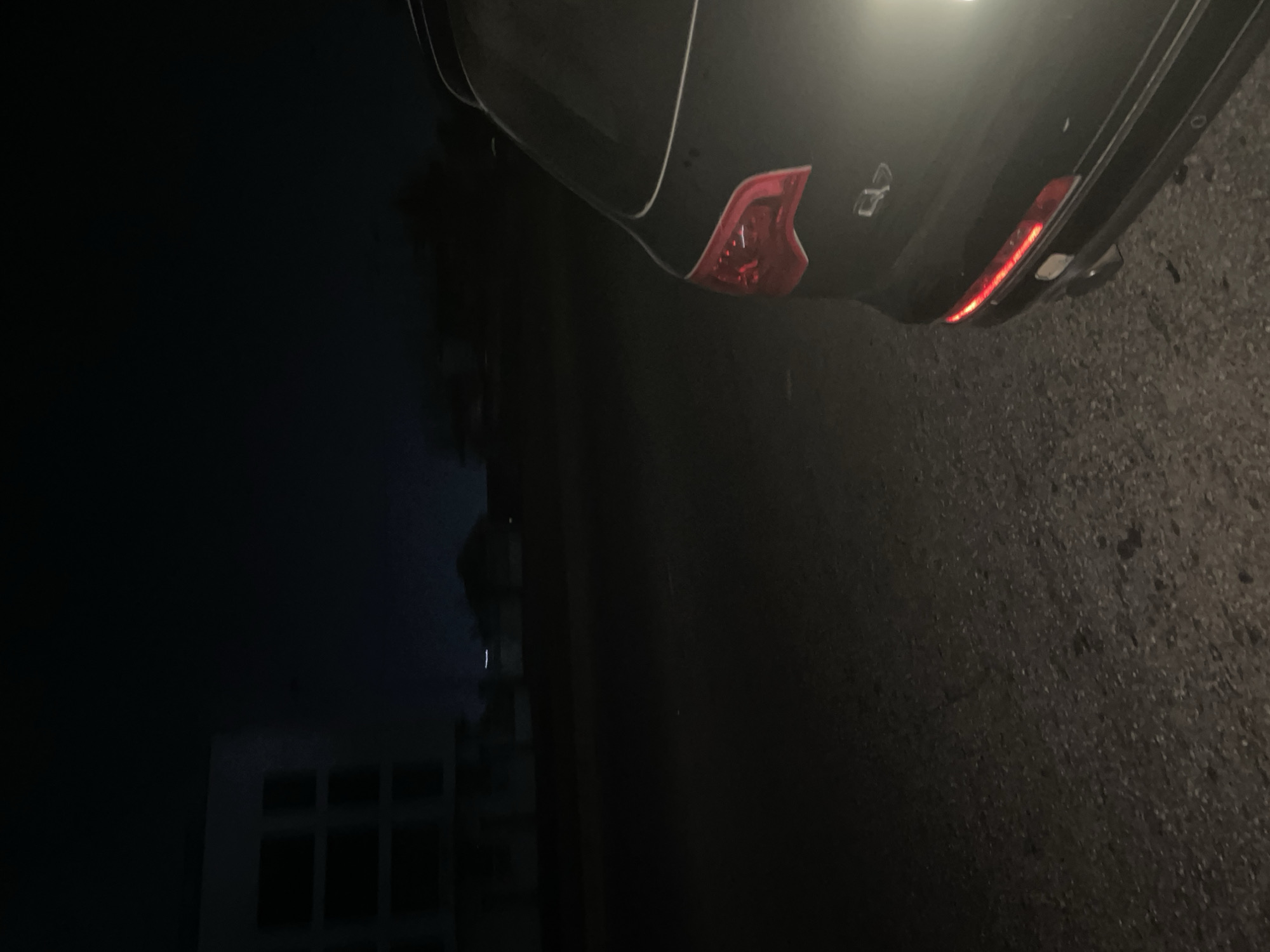 A night scene in Dutsen Alhaji, Nigeria shows the rear of a black car on an asphalt surface. The car's tail lights are illuminated. In the background, a building with several windows is visible. The lighting is dim.