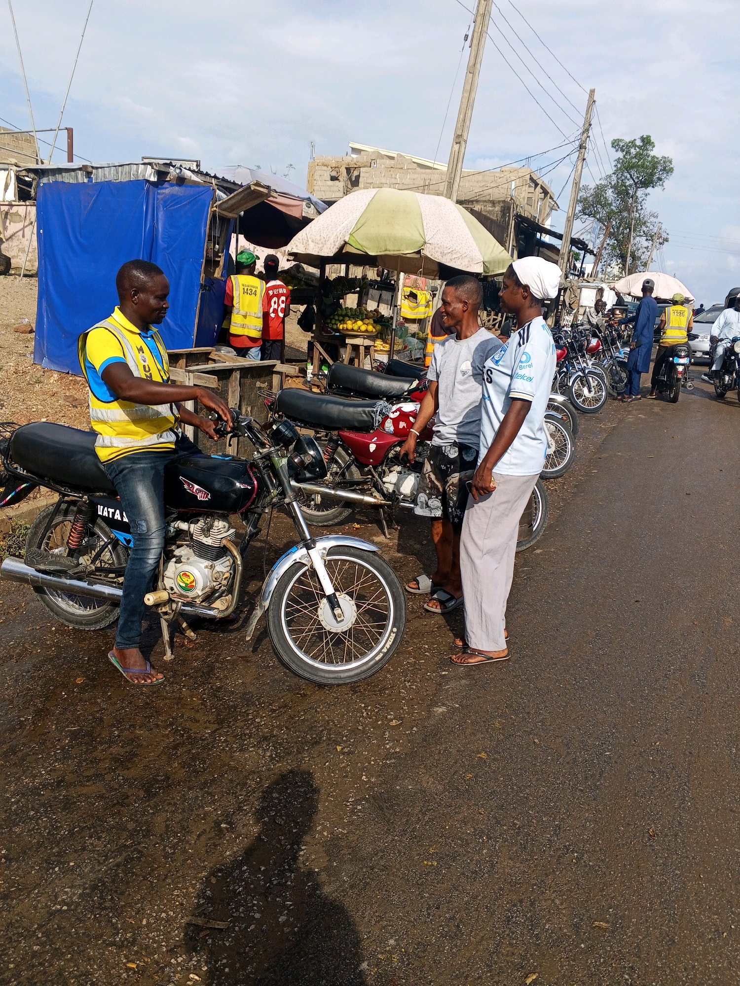 In Zariagi, Nigeria, on a sunny day, a man wearing a yellow and blue reflective vest and dark jeans sits on a black motorcycle. Another man in a grey t-shirt and patterned shorts stands to his right, talking with a third man who is wearing a light blue t-shirt and light grey pants. Behind them, more motorcycles are parked, some with riders. A fruit stand with oranges and bananas is visible under a green and white umbrella. In the background, buildings with corrugated metal roofs and cinder block walls suggest a market or informal settlement. Overhead, electrical wires are strung between wooden poles. The ground is a mix of dirt and asphalt, with some wet patches reflecting the sunlight. The scene depicts a lively roadside gathering, possibly a transport hub or market area, with people engaged in conversation and activity.