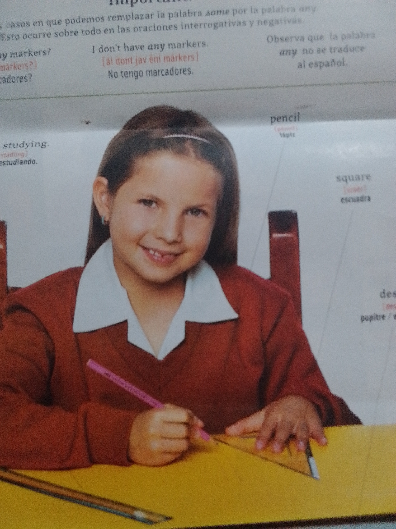 This media file is a photograph of a young girl in what appears to be a classroom setting. The girl, who is the primary subject, is wearing a red v-neck sweater over a white collared shirt and has her hair pulled back. She is smiling at the camera and holding a pink pencil, using a ruler and a triangle ruler on a yellow surface. The background shows educational materials with text in both English and Spanish.
The visible text includes phrases like "casos en que podemos remplazar la palabra some por la palabra any," "Esto ocurre sobre todo en las oraciones interrogativas y negativas," "I don't have any markers," and "No tengo marcadores." Other labels like "pencil," "square," and "des" with their Spanish translations "lápiz," "escuadra," and "pupitre/e" respectively, are also present, suggesting a lesson on vocabulary or grammar.
The scene is brightly lit, indicating it could be daytime. The girl's expression is happy and engaged. The setting appears to be a school in La Tagua, Venezuela, given the context provided. The focus is on the girl and the learning materials around her, suggesting an educational activity.