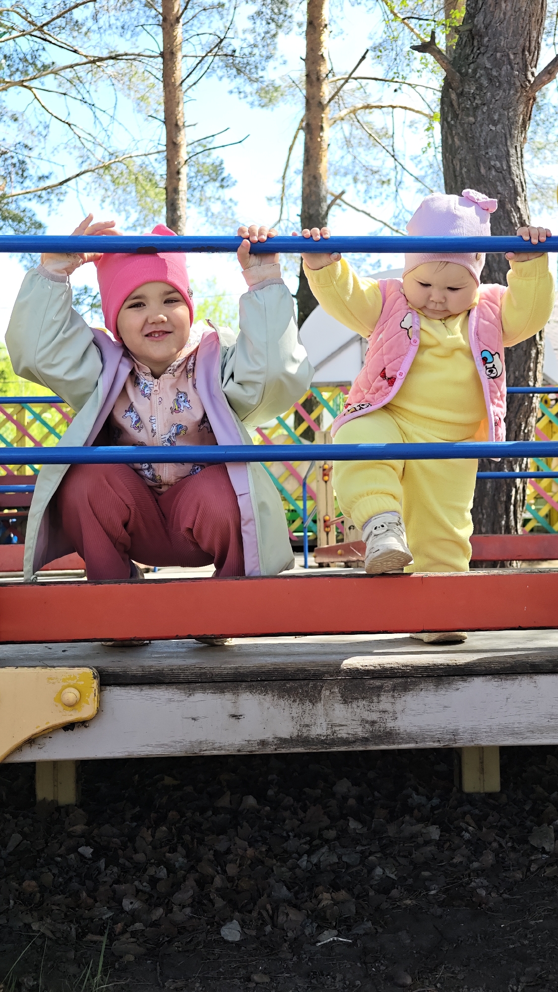 Two young children are on a playground structure with blue metal bars and a wooden platform. The child on the left is wearing a pink hat, a jacket with a unicorn pattern, maroon pants, and a light green coat. She is smiling and squatting, holding onto the blue bar with both hands. The child on the right is wearing a light pink hat with a bow, a yellow outfit, and a pink vest with a dog pattern. She is standing, holding the blue bar with both hands, and wearing white shoes. Behind the children, there is a tall tree and a colorful wooden fence. The ground beneath the playground structure is covered with dark leaves and debris. The location is Chaikovsky, Russia.