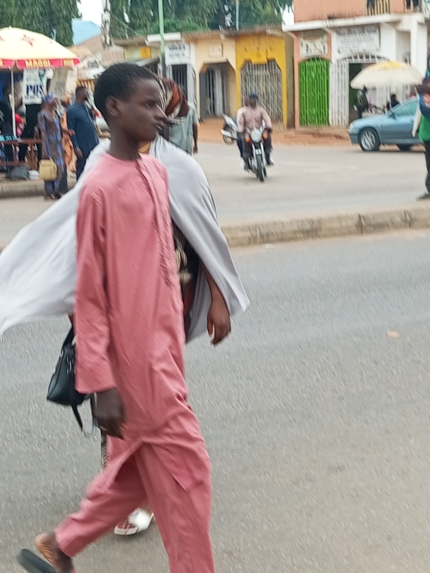 The image shows a street scene in Gombe, Nigeria. In the foreground, a young boy in a pink tunic and pants walks towards the right of the frame. He is carrying a black bag slung over his shoulder. Behind him, a person in a grey robe is visible. The background features a street with several buildings, a blue car, and a motorcycle with a rider. People are seen walking and standing on the sidewalk. The scene appears to be a typical urban environment with market stalls and shops.